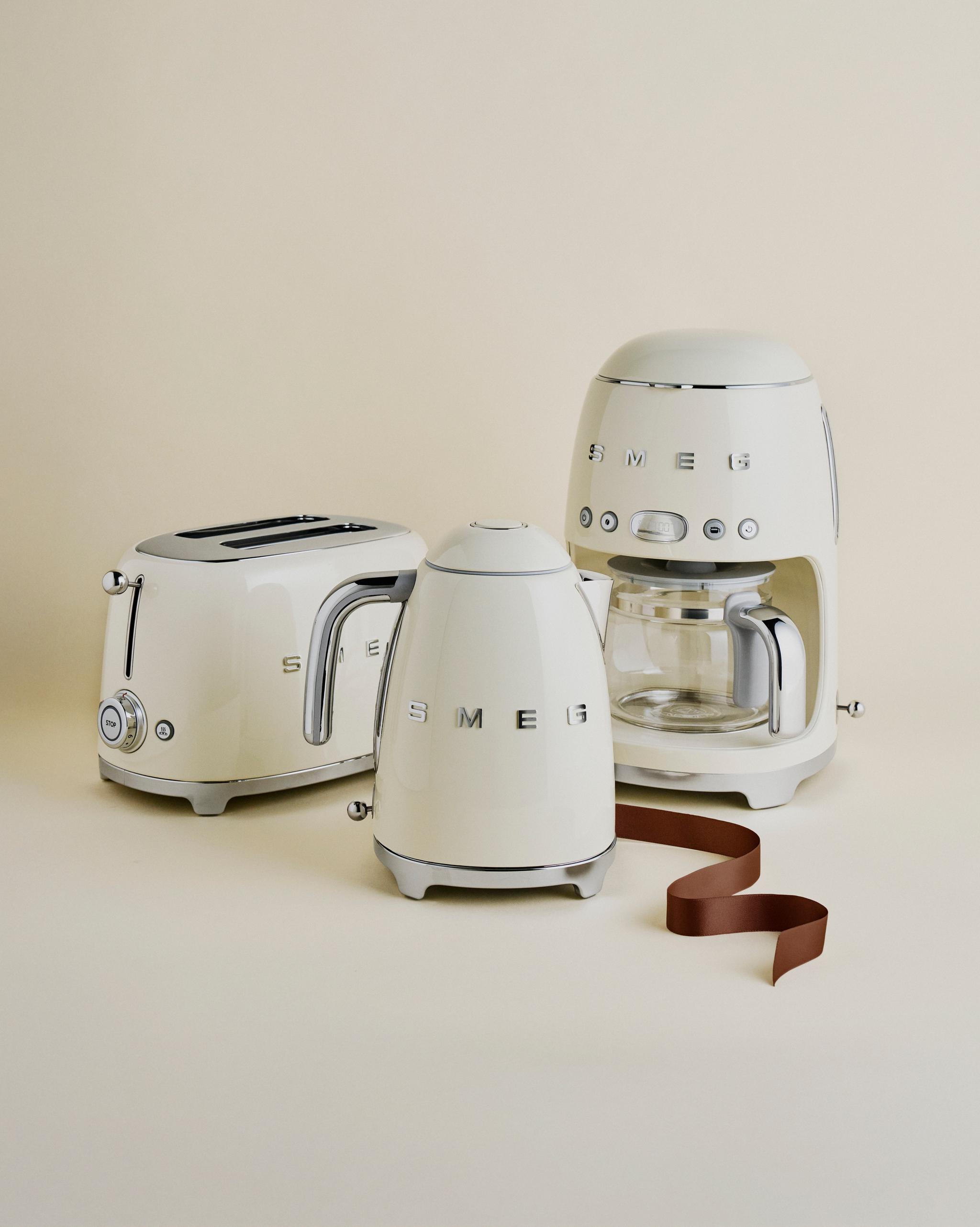 Kremfargede Smeg kjøkkenapparater – brødrister, vannkoker og kaffetrakter – står stilrent oppstilt mot en lys beige bakgrunn.