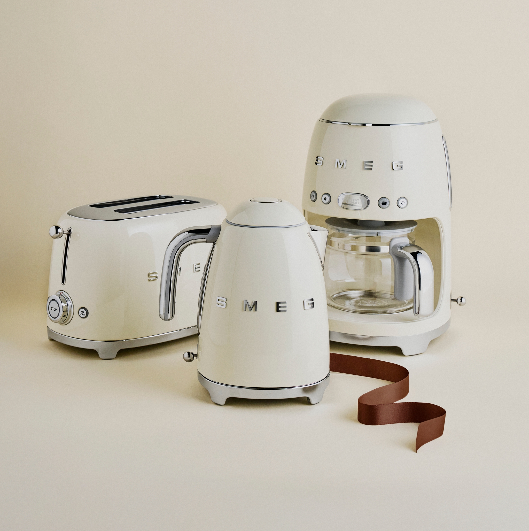Kremfargede Smeg kjøkkenapparater – brødrister, vannkoker og kaffetrakter – står stilrent oppstilt mot en lys beige bakgrunn.