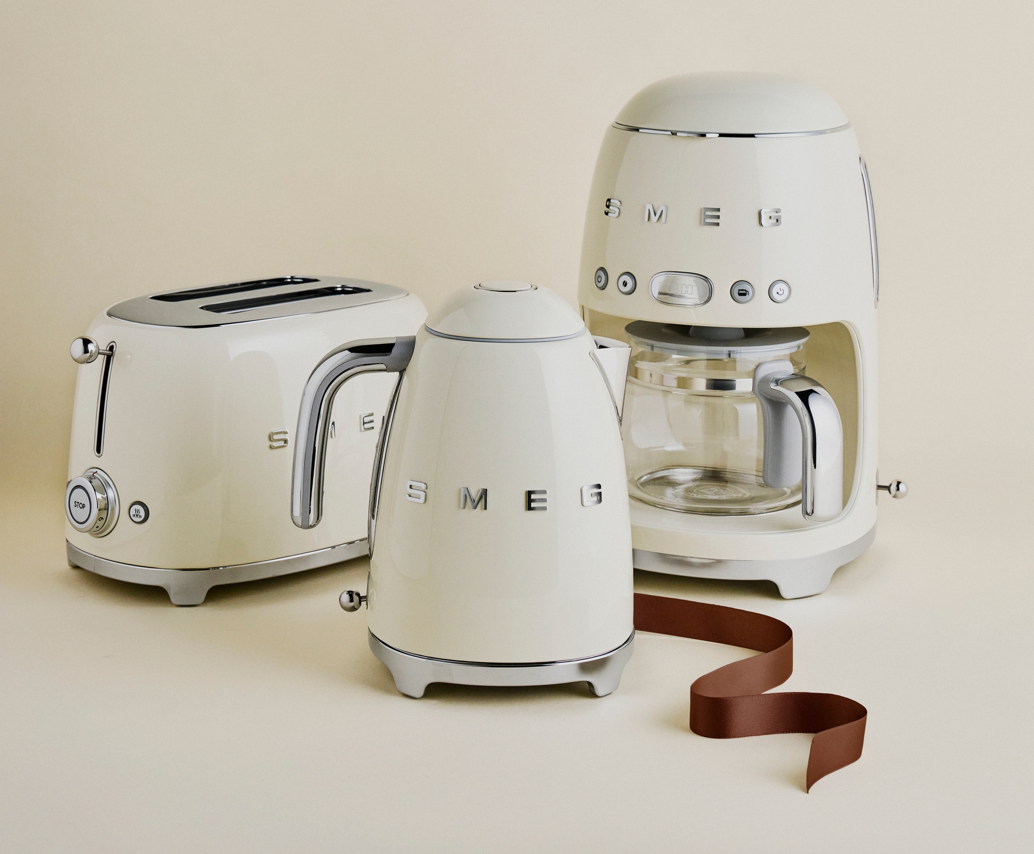 Krämfärgade SMEG köksapparater – brödrost, vattenkokare och kaffebryggare – står uppställda mot en beige bakgrund.