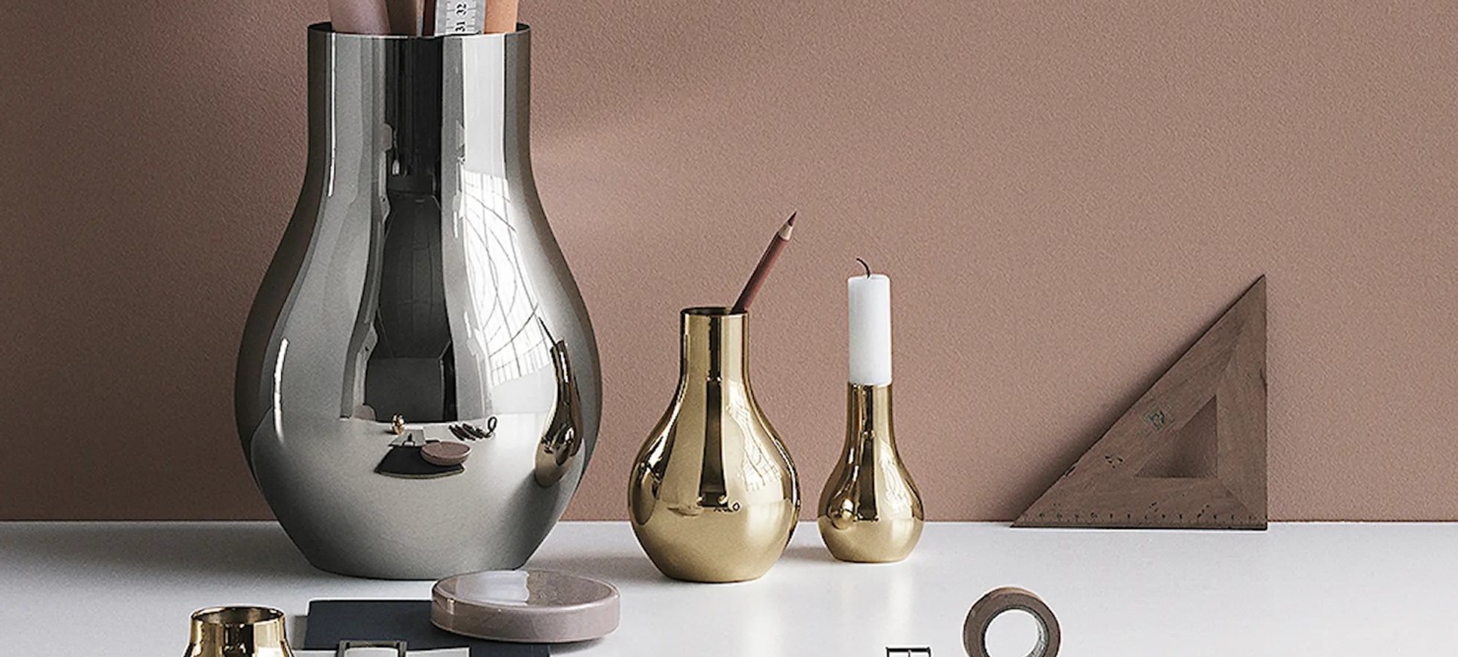 Stilleben med moderne metallvaser, lysestake og skrivebordstilbehør mot en beige vegg.