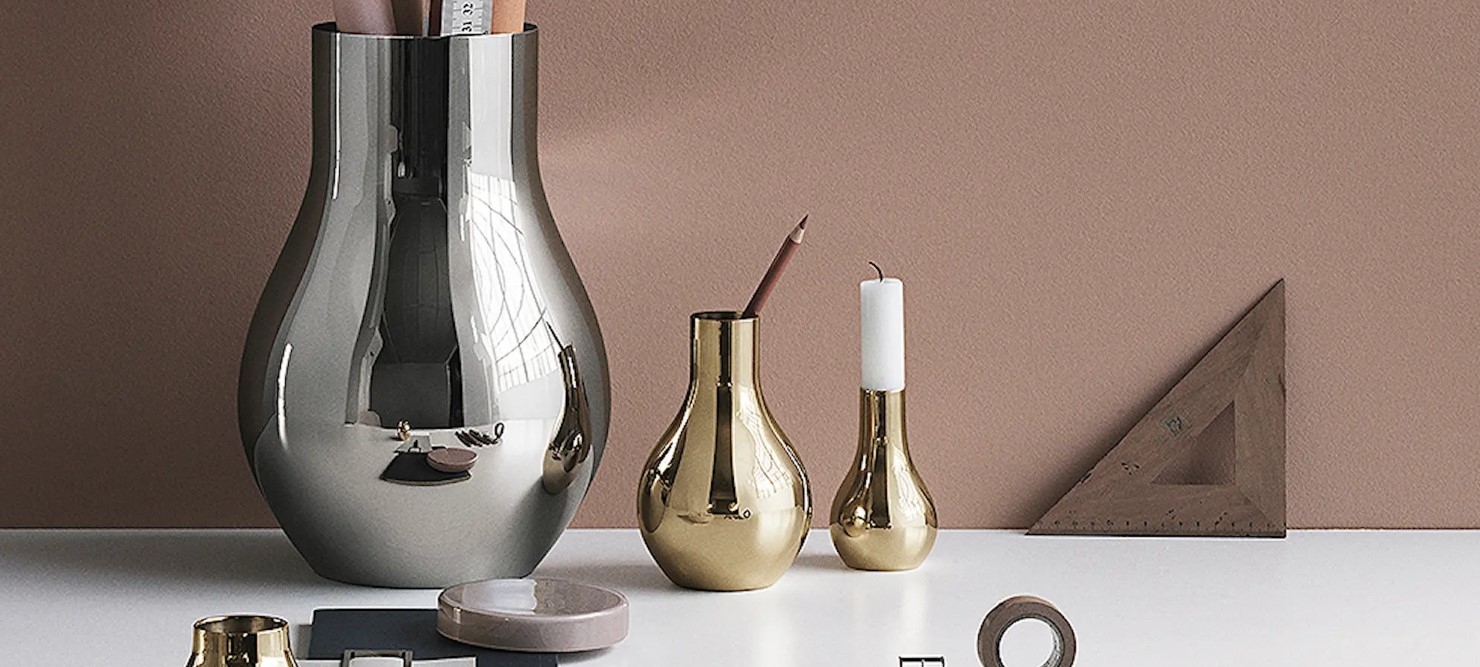 Stilleben med moderne metallvaser, lysestake og skrivebordstilbehør mot en beige vegg.