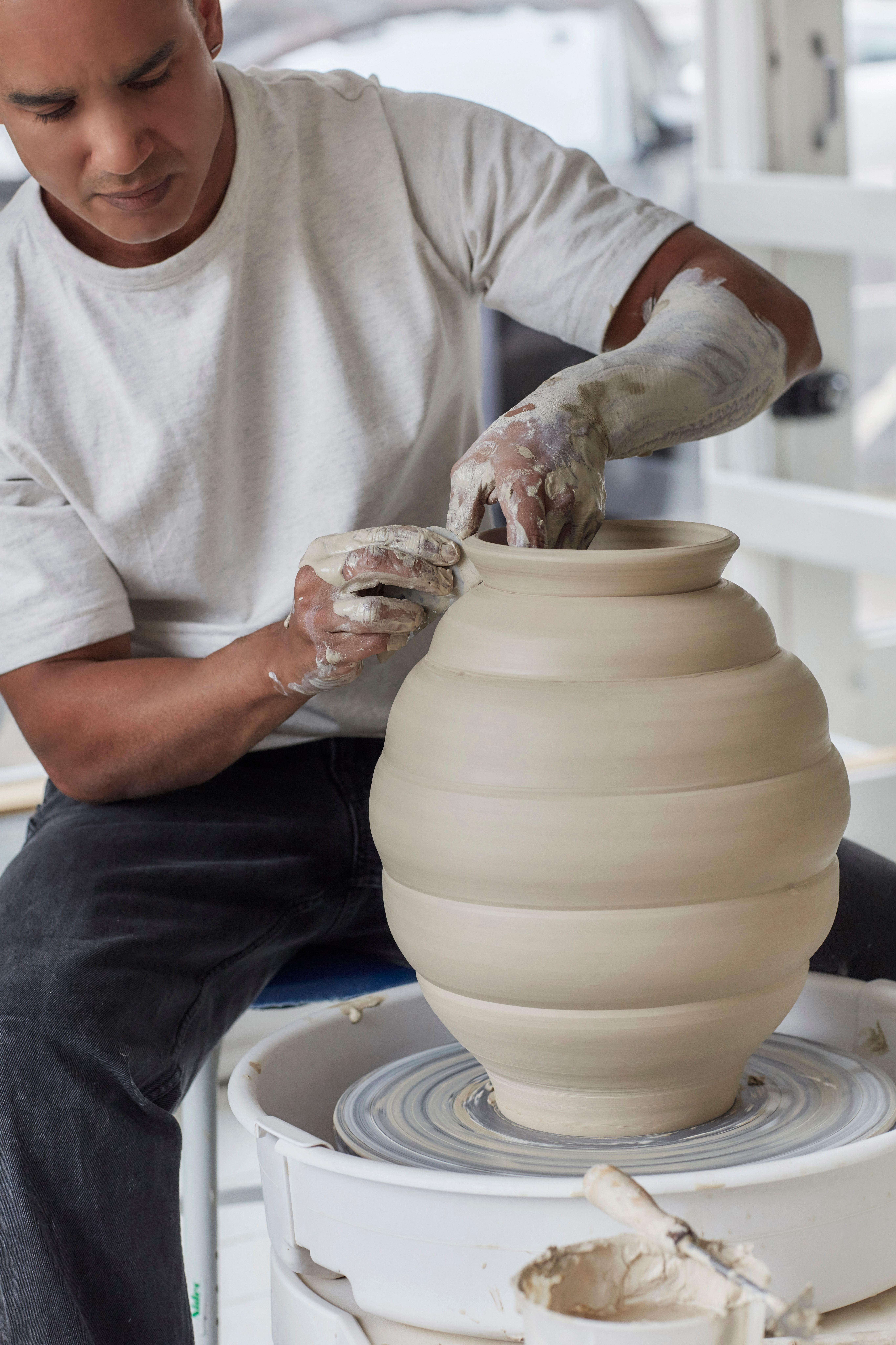 A person with clay-covered hands shapes a large, ribbed clay pot on a pottery wheel.