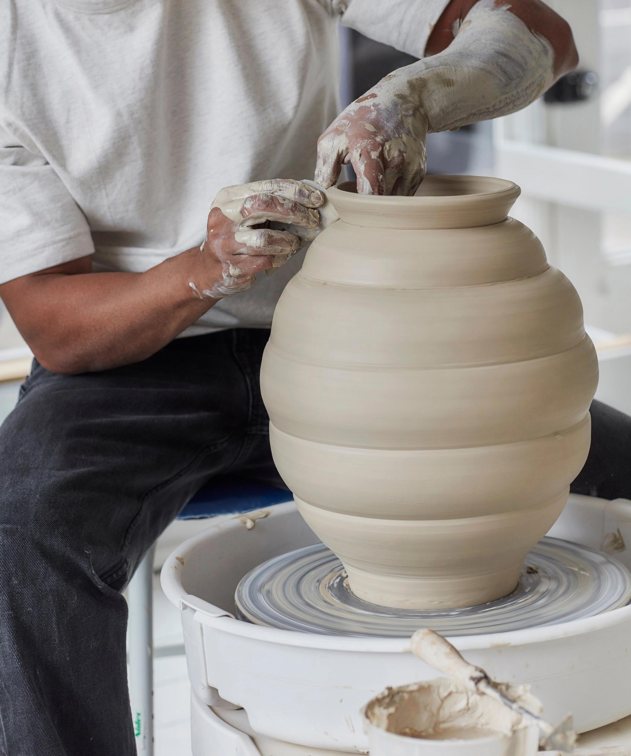 A person with clay-covered hands shapes a large, ribbed clay pot on a pottery wheel.