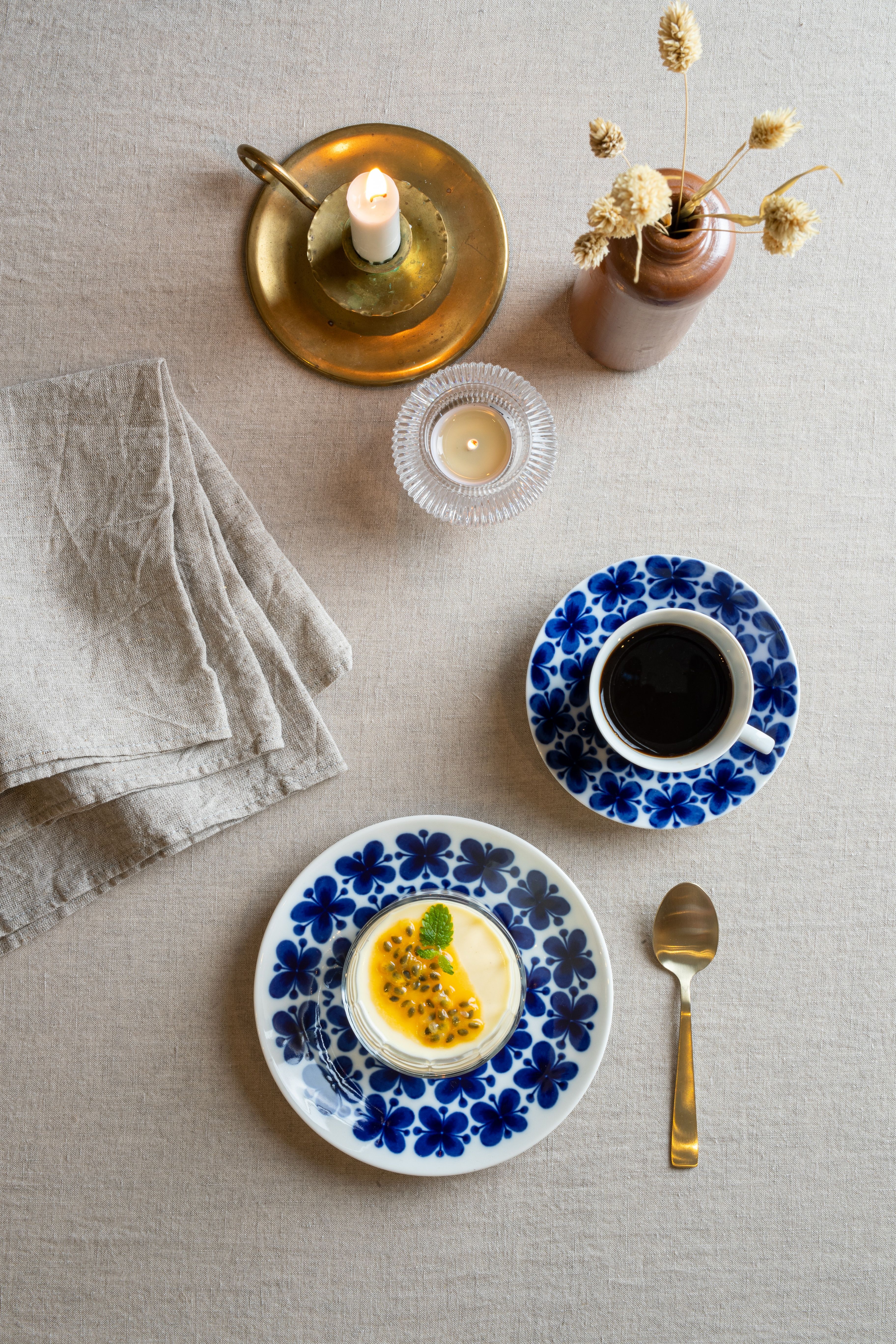 Dukat fikabord med kaffe, passionsfruktsdessert, levande ljus och blåblommigt porslin på en linneduk.