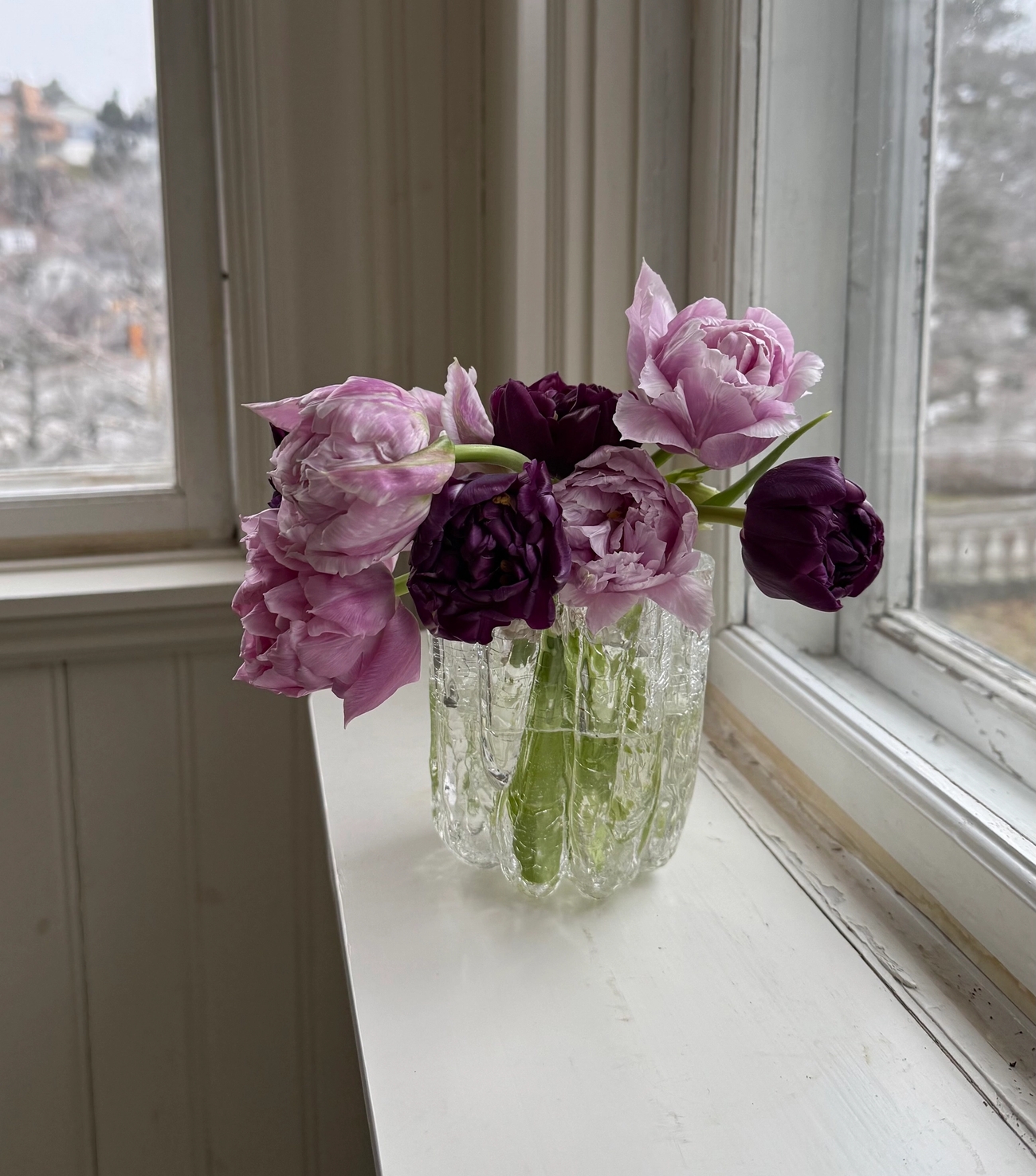 En bukett lila och ljusrosa tulpaner i en texturerad glasvas på en vit fönsterbräda.