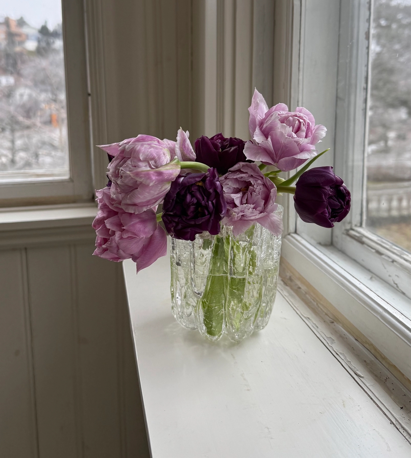 En bukett lila och ljusrosa tulpaner i en texturerad glasvas på en vit fönsterbräda.