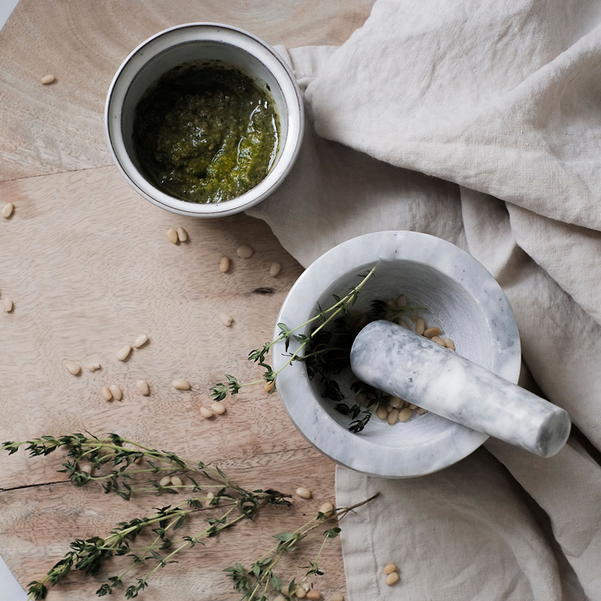 En morter og støter står på et trebord ved siden av en skål med pesto