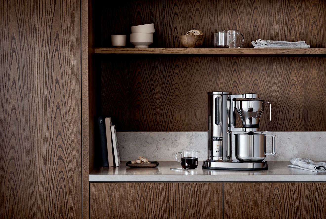 A modern silver coffee maker on a light marble counter with dark wood cabinets and shelves.
