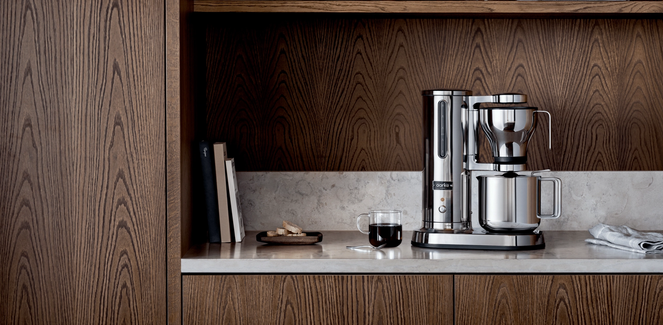 A modern silver coffee maker on a light marble counter with dark wood cabinets and shelves.