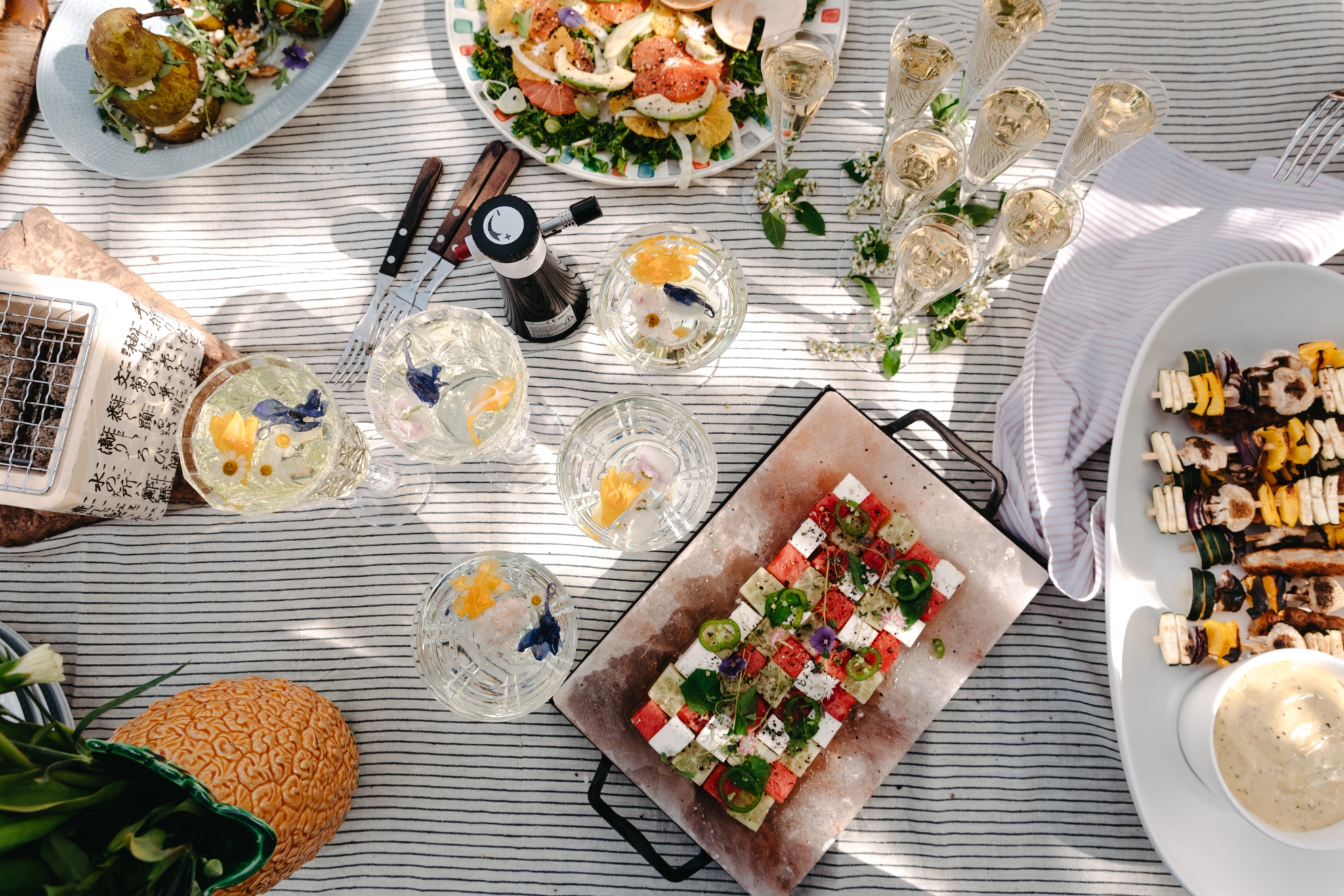 Dekket bord ovenfra med feta- og vannmelonspyd, glass, grillspyd og salat på en stripete duk