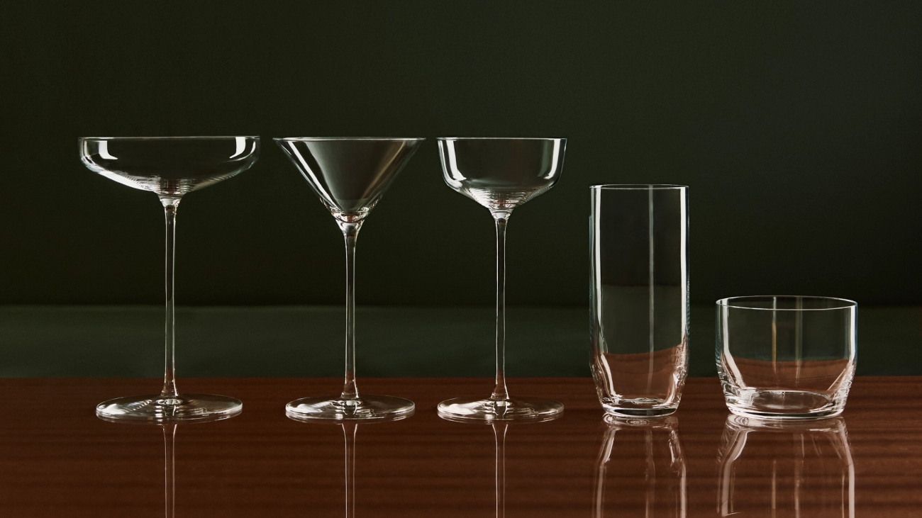 Five clear glass drinkware items: two coupe glasses, a martini glass, a highball glass, and a rocks glass, on a wooden table.Fem barglas i serien Orrefors x Björn Frantzén mot en mörk bakgrund