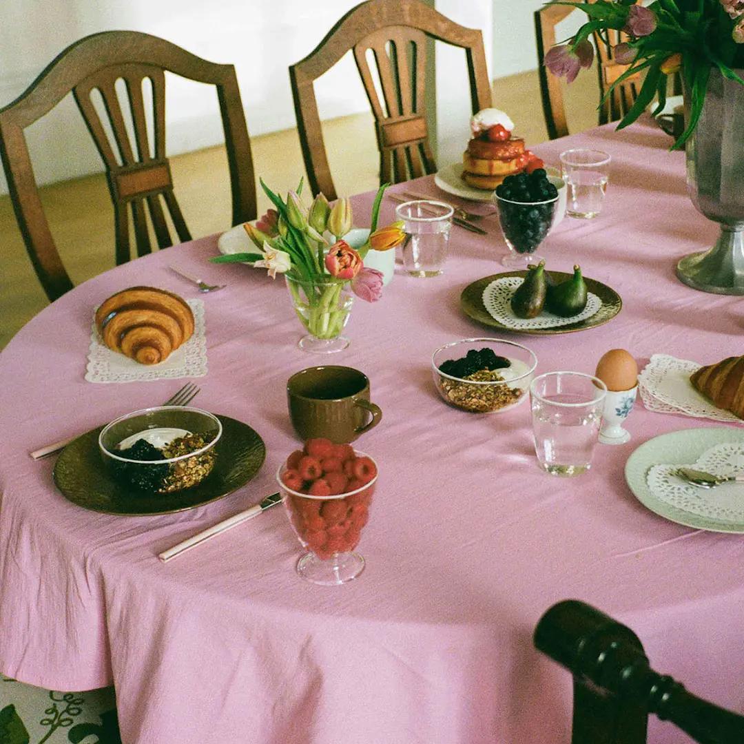 A pink-covered table laden with a colorful breakfast spread including croissants, pancakes, fresh fruit, yogurt, and flowers.