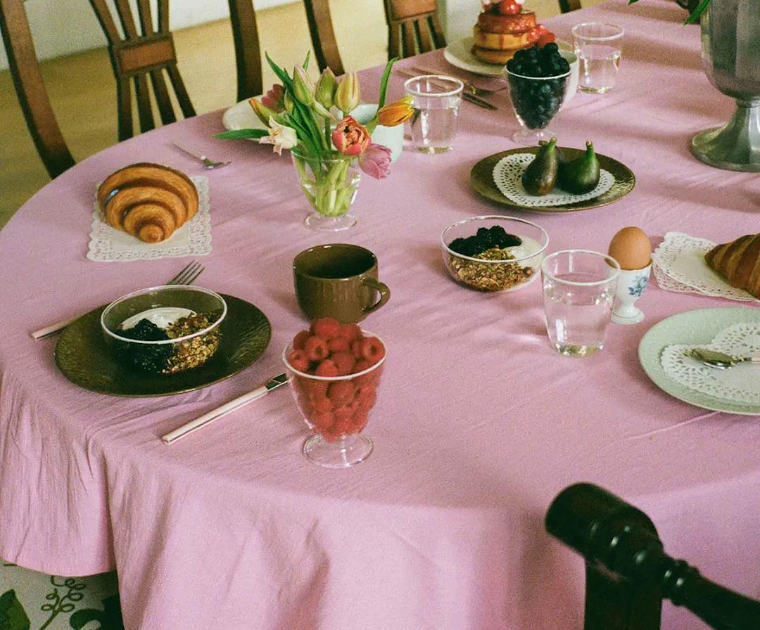 A pink-covered table laden with a colorful breakfast spread including croissants, pancakes, fresh fruit, yogurt, and flowers.