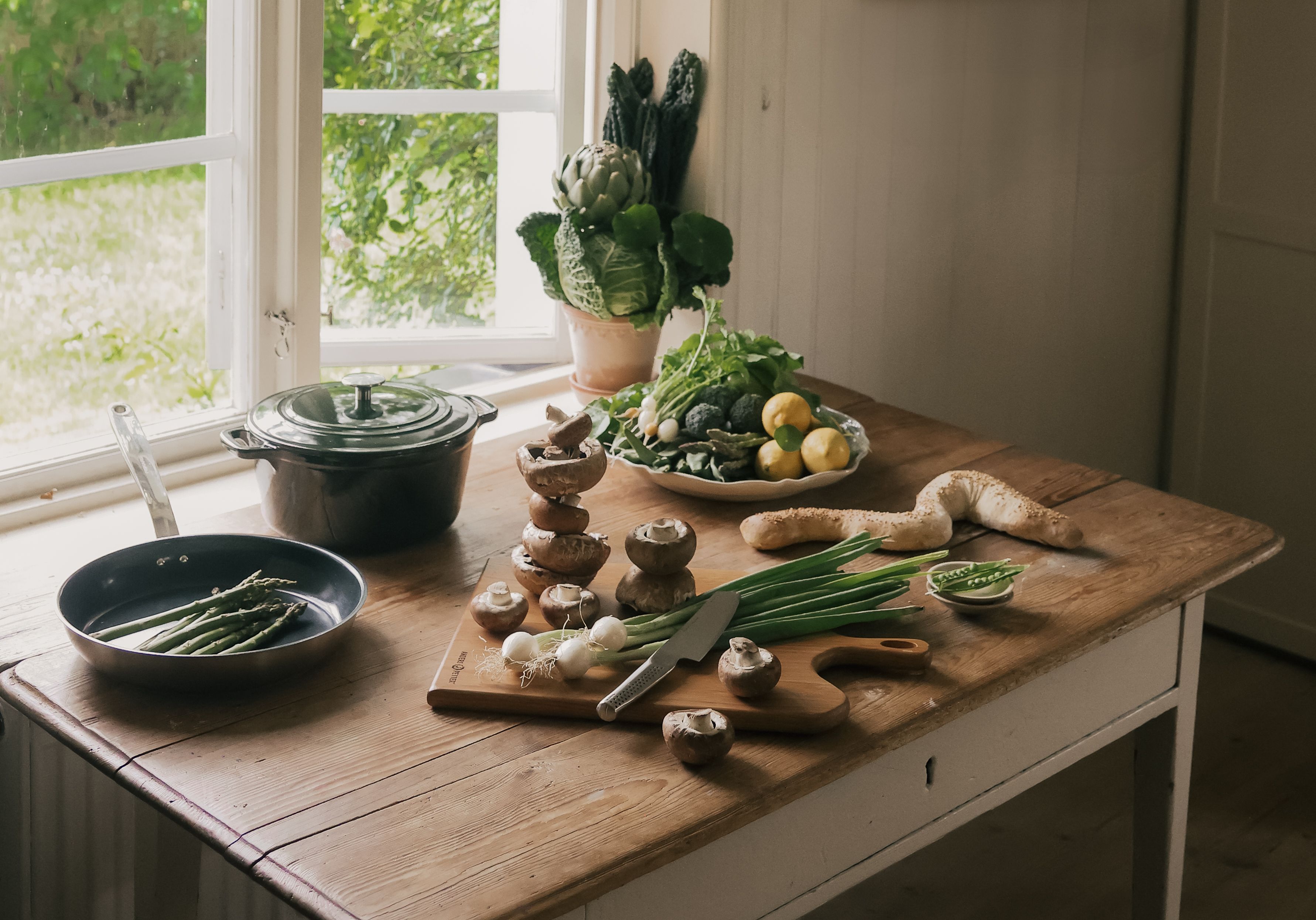 Rustikt köksbord med färska grönsaker som sparris, staplade svampar och grönsaker, bredvid kastruller vid ett ljust fönster.