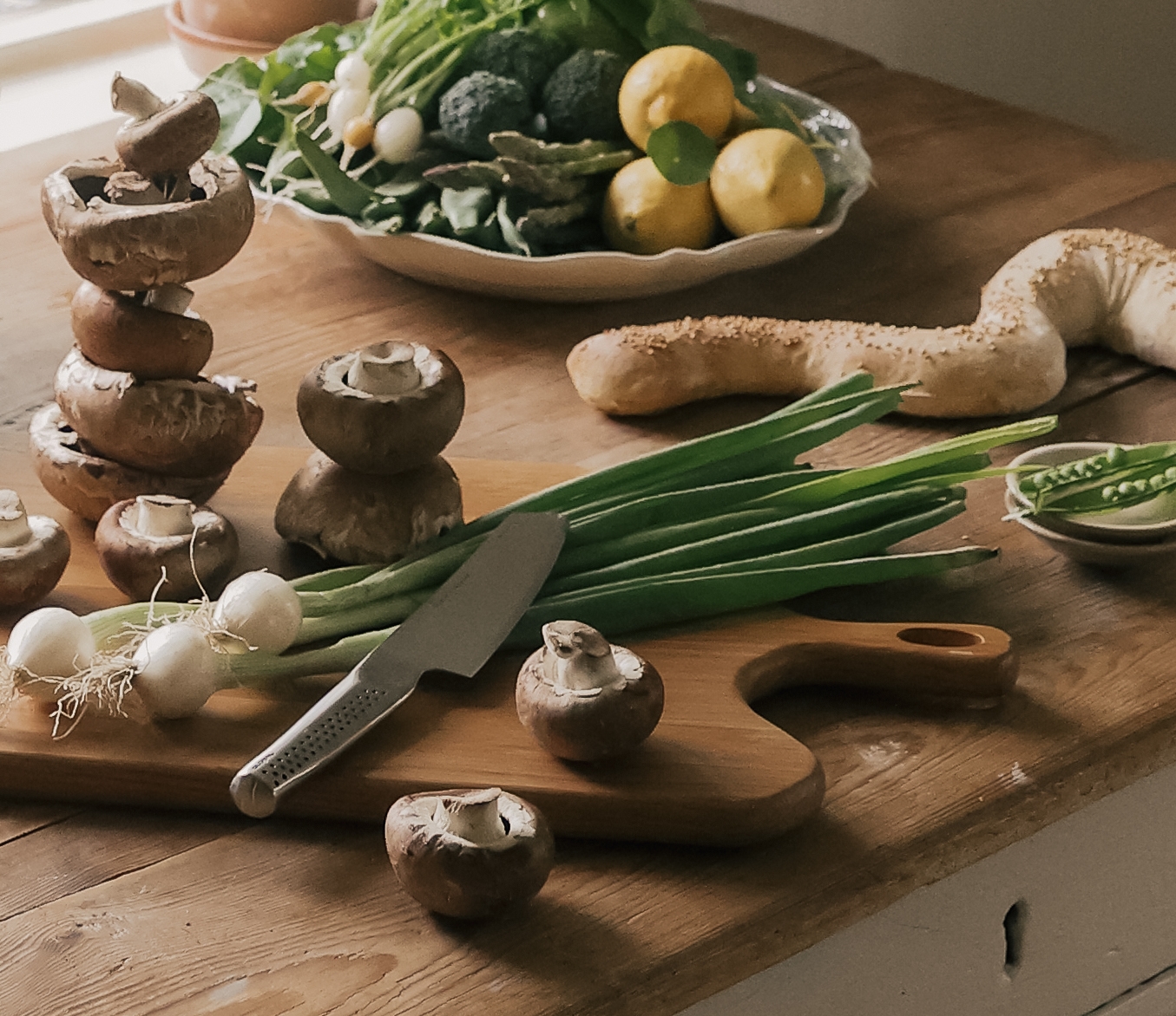 Rustikt köksbord vid ett fönster dukat med färska grönsaker, svamp, bröd och köksredskap redo för matlagning.