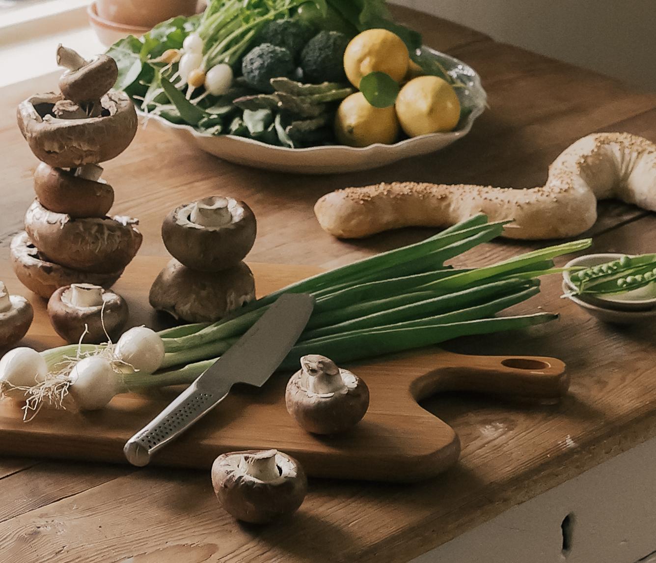 Rustikt köksbord vid ett fönster dukat med färska grönsaker, svamp, bröd och köksredskap redo för matlagning.