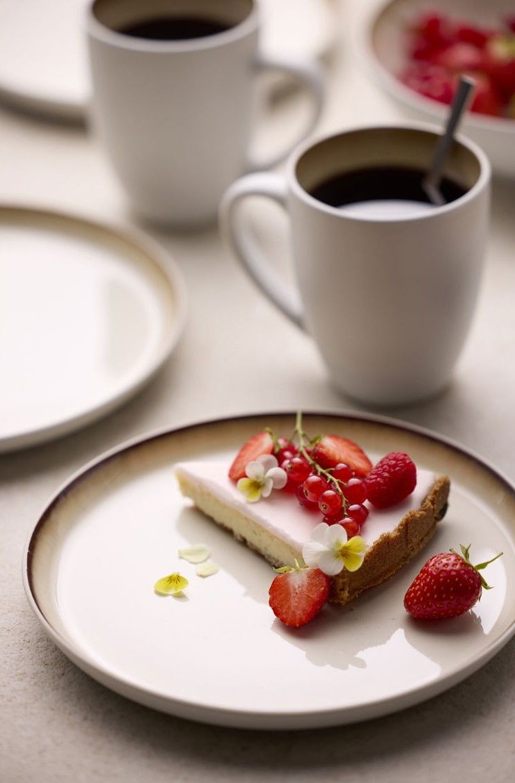 A slice of cheesecake topped with strawberries, red currants, and edible flowers on a plate, with two mugs of coffee in the blurred background.