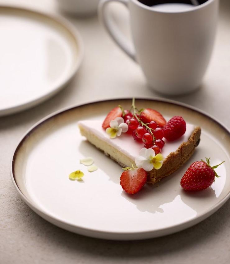 A slice of cheesecake topped with strawberries, red currants, and edible flowers on a plate, with two mugs of coffee in the blurred background.