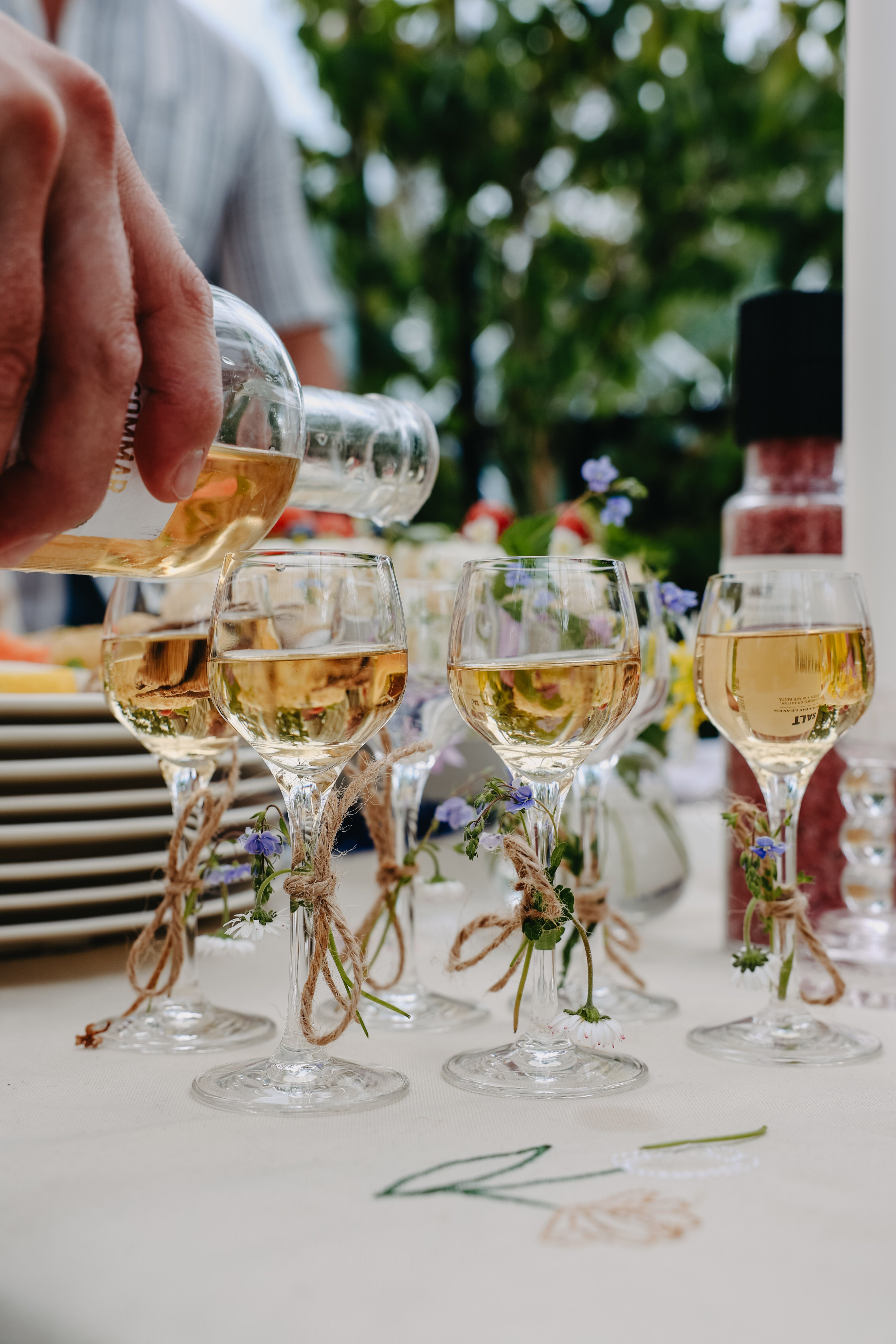 Närbild på en hand som häller vitt vin i dekorerade vinglas på ett somrigt dukat bord utomhus.