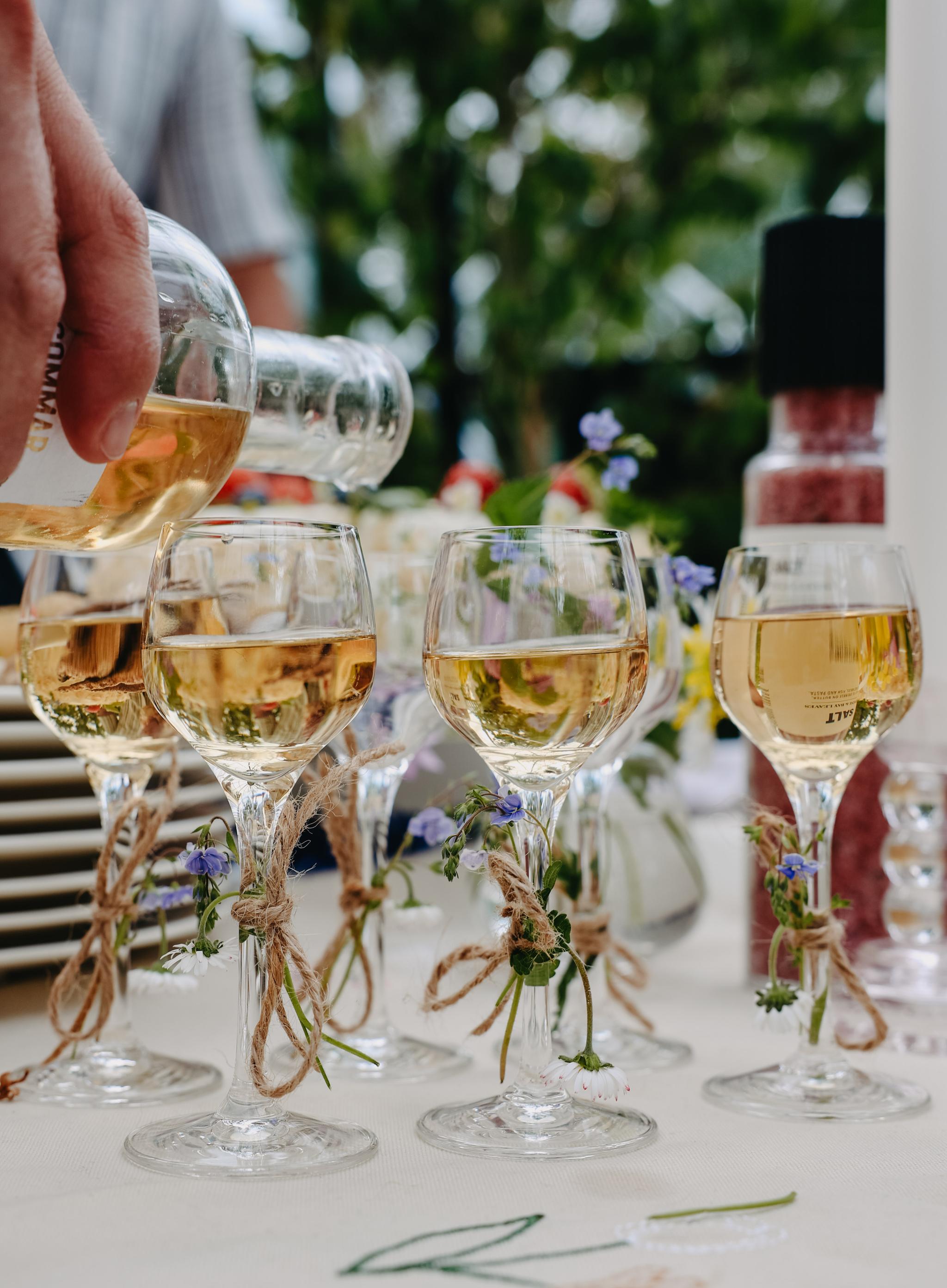 Närbild på en hand som häller vitt vin i dekorerade vinglas på ett somrigt dukat bord utomhus.