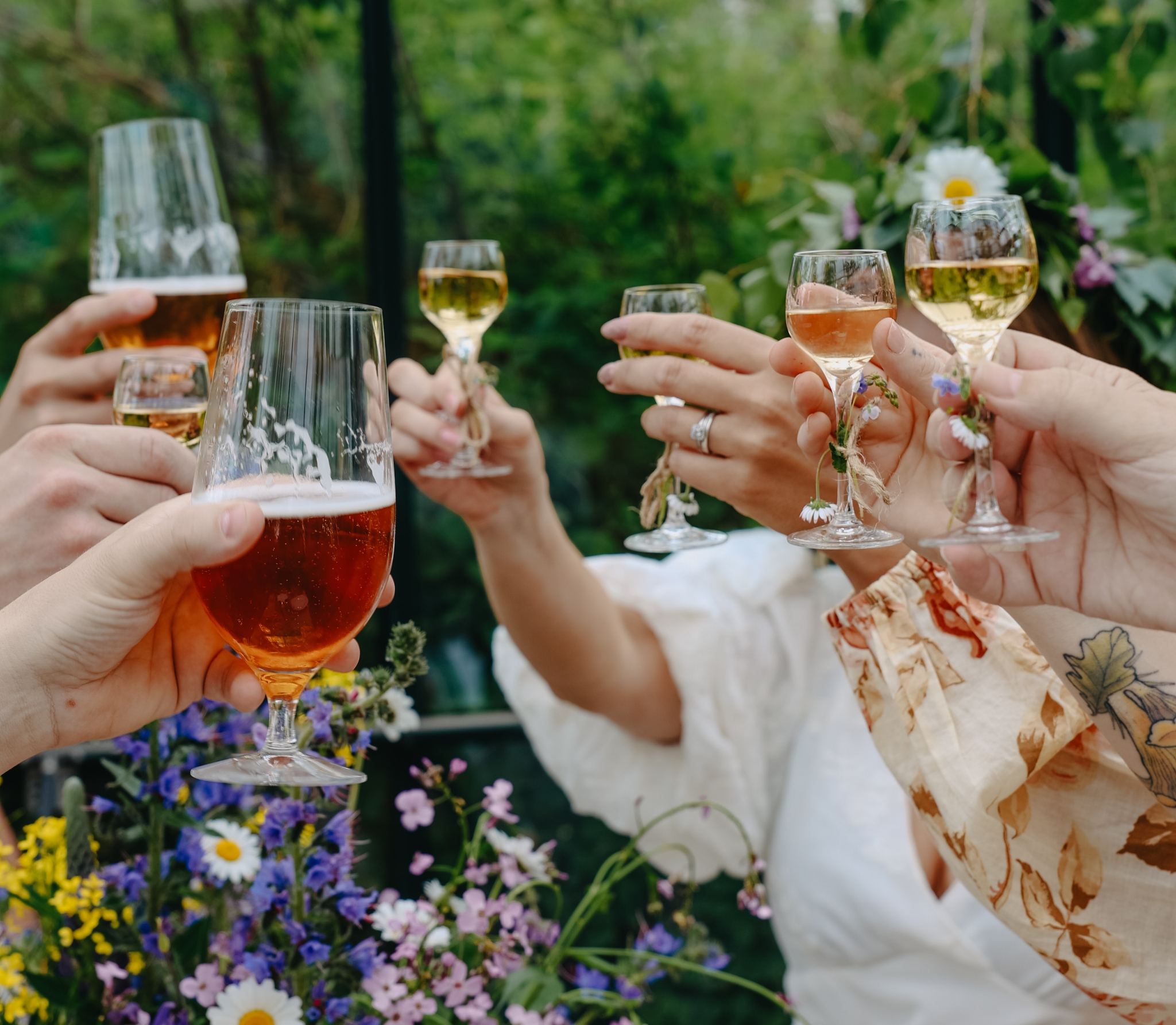 Vänner skålar med öl och vin bland sommarblommor under en grönskande svensk midsommarfest.