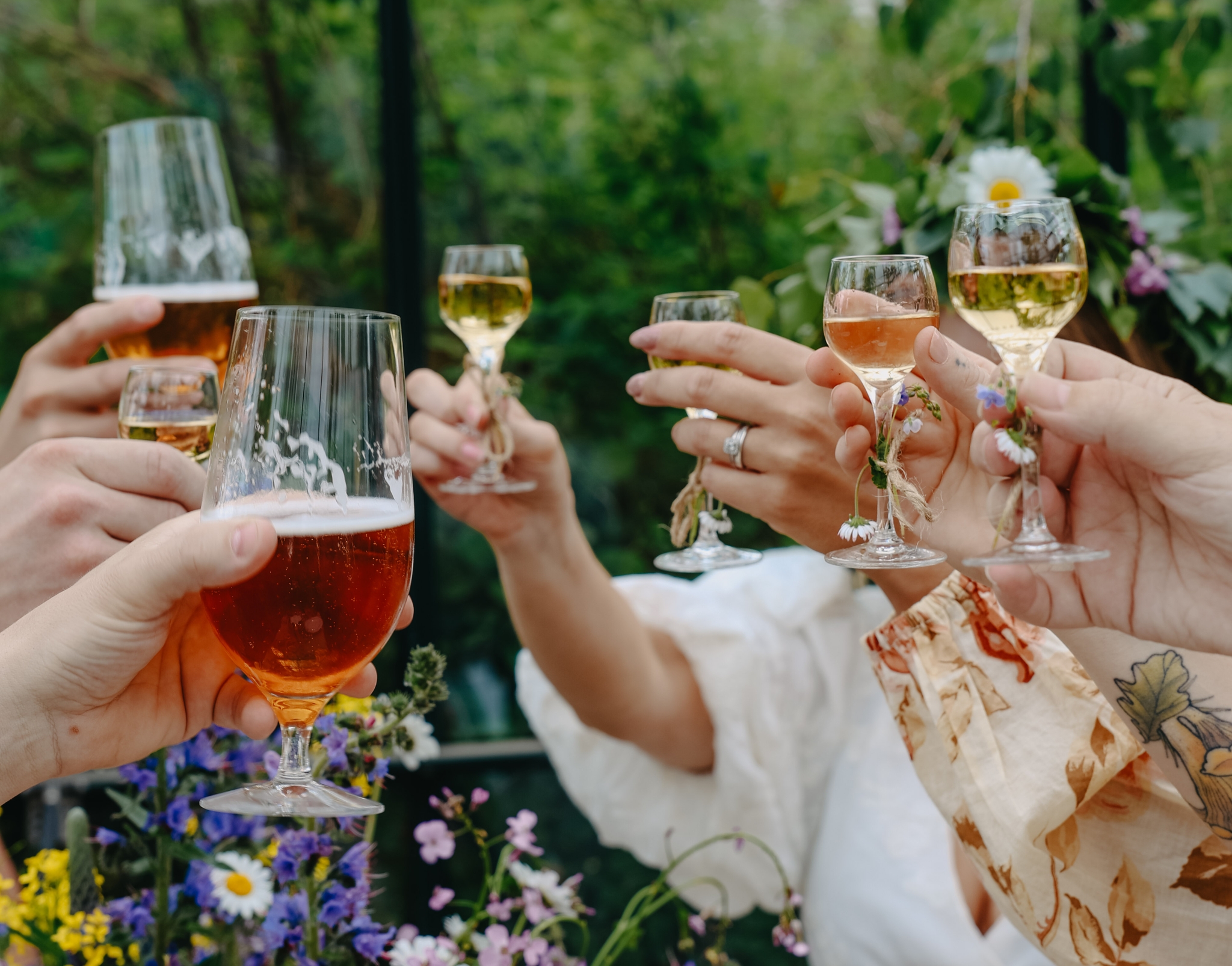 Vänner skålar med öl och vin under en somrig svensk fest omgiven av gröna blad och blommor.