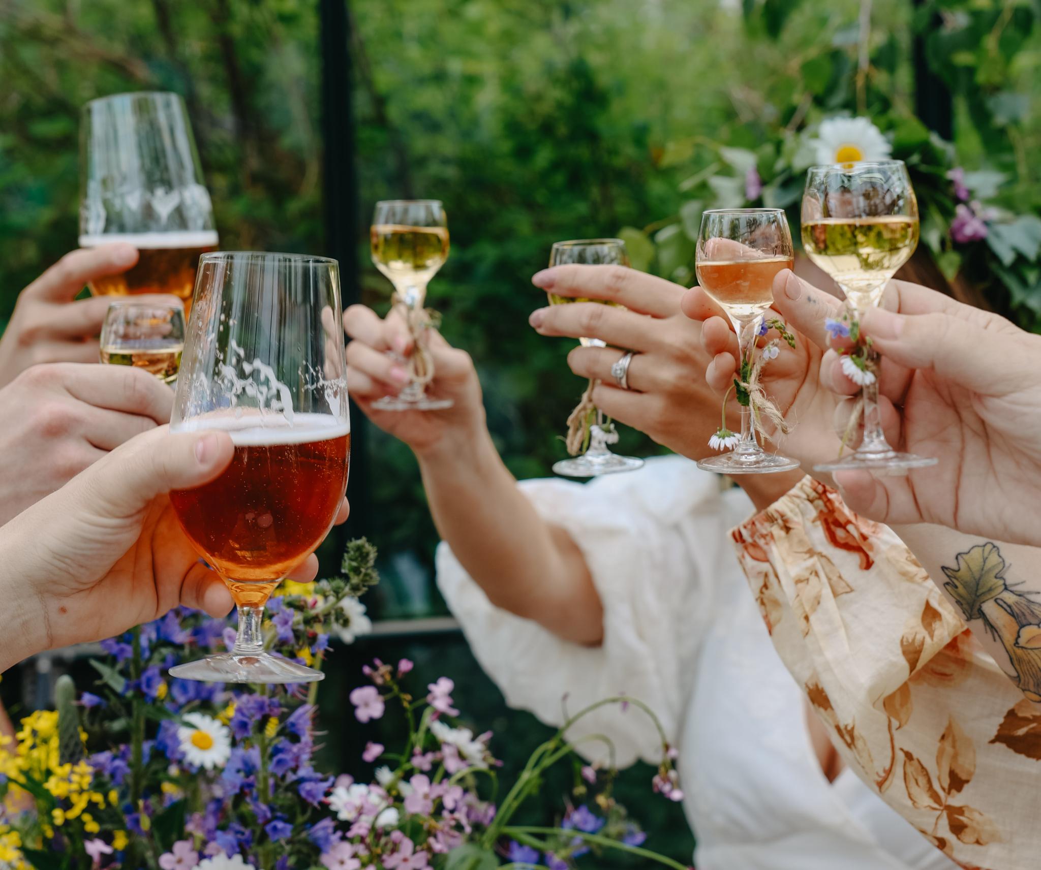 Venner skåler med øl og vin i en grønn hage omgitt av sommerblomster.