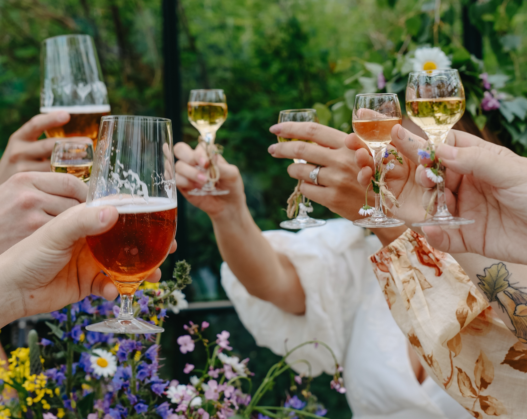 Venner skåler med øl og vin i en grønn hage omgitt av sommerblomster.
