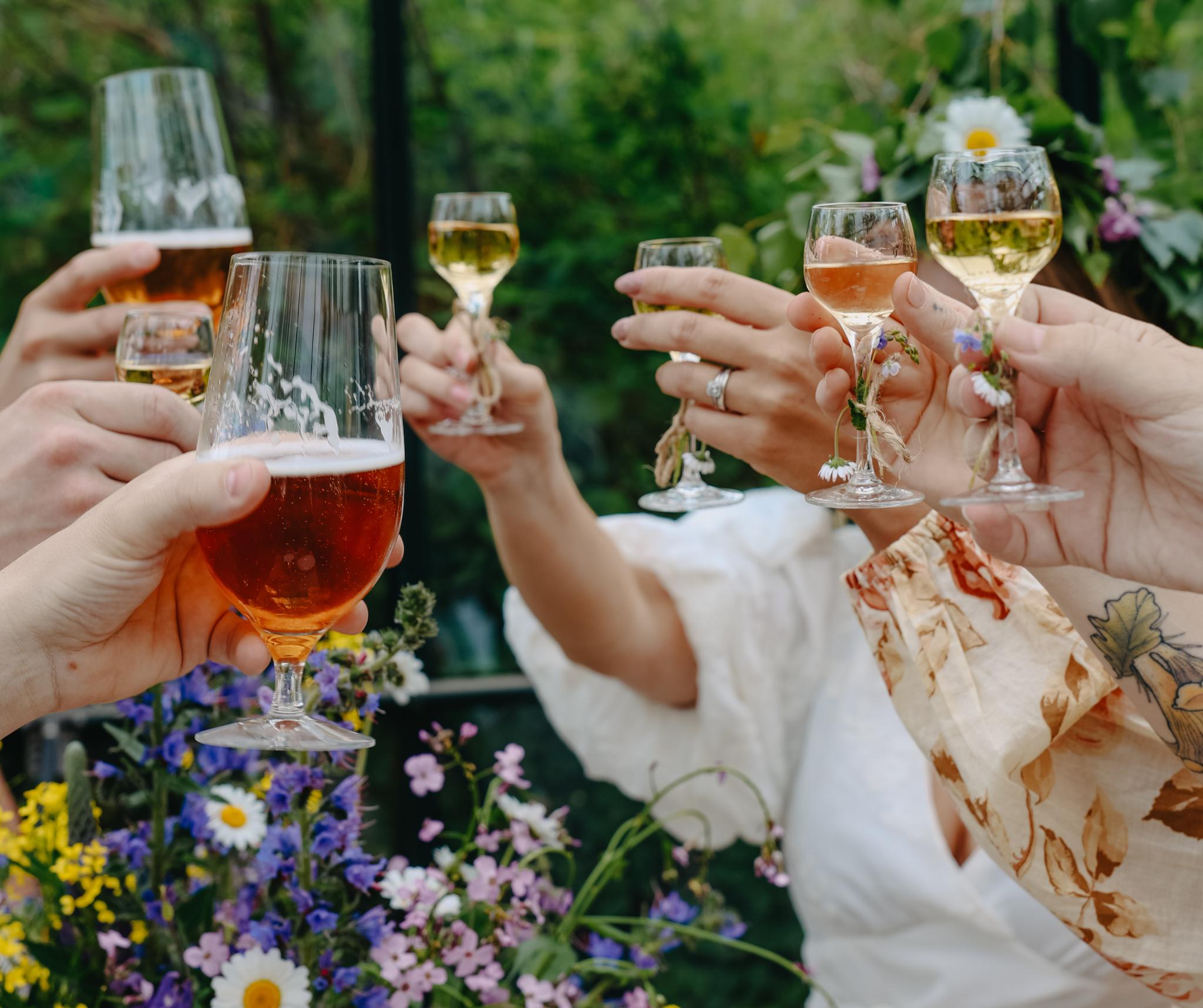 Vänner skålar med öl och vin bland sommarblommor under en grönskande svensk midsommarfest.