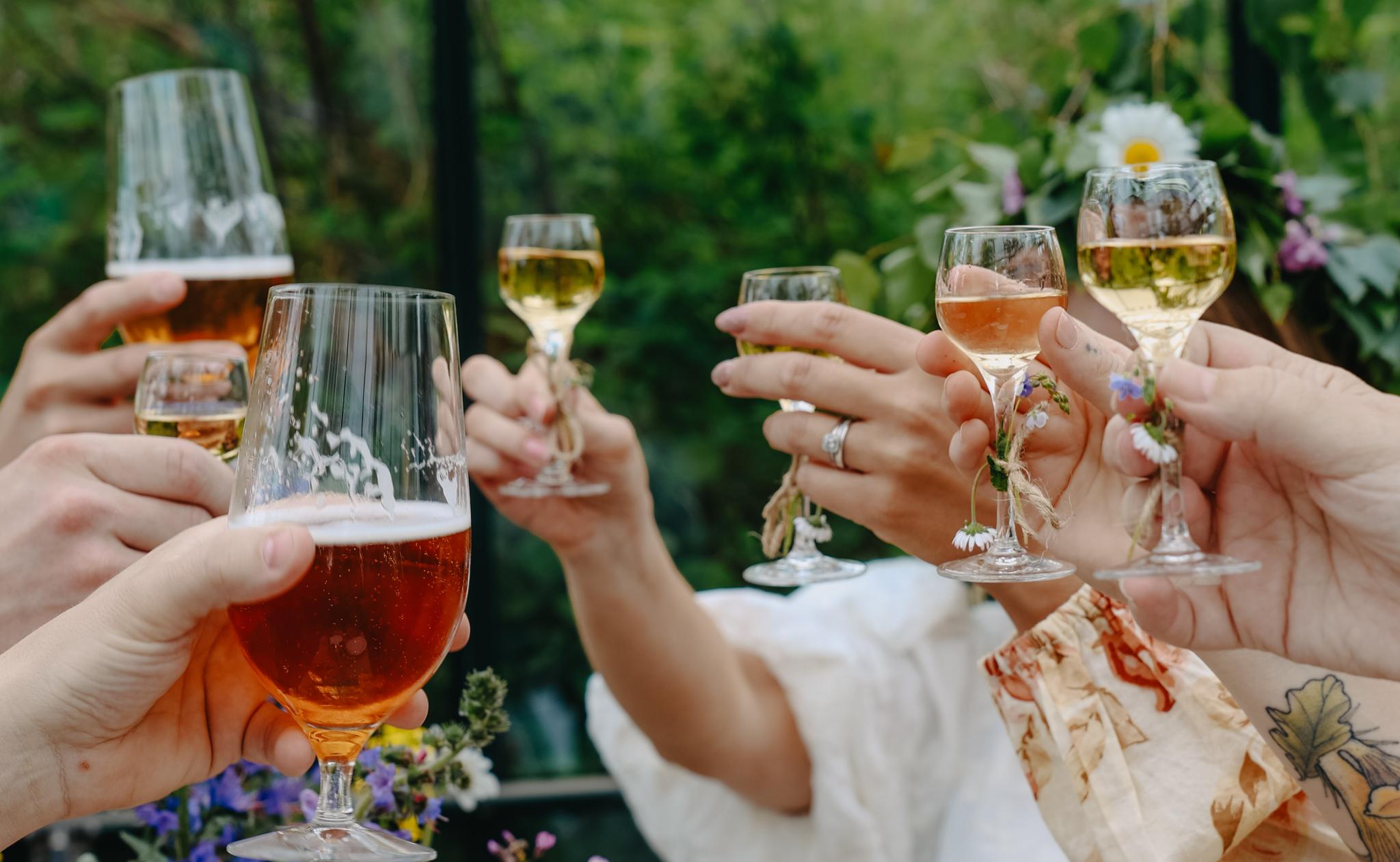 Vänner skålar med öl och vin bland sommarblommor under en grönskande svensk utomhusfest.