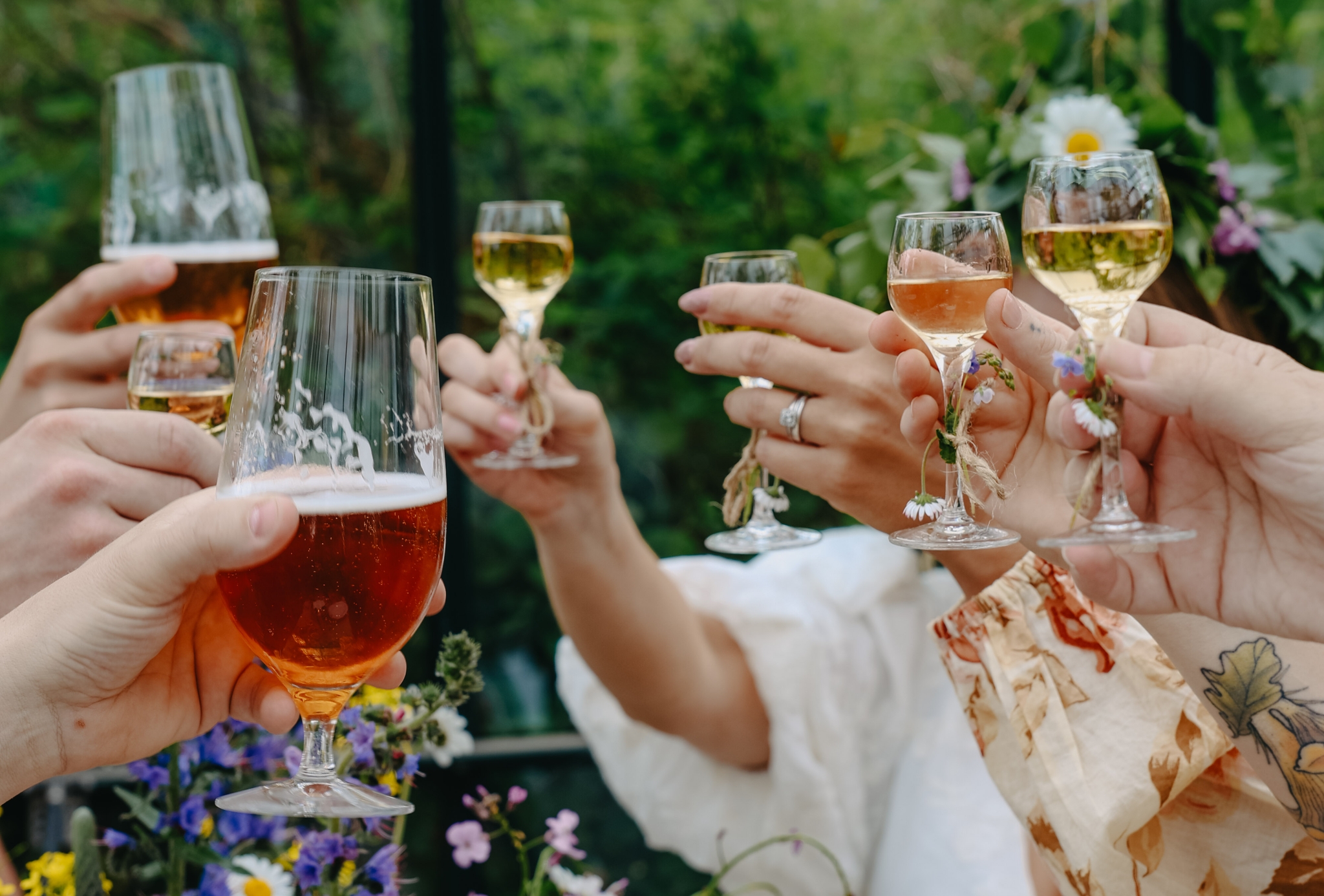 Venner skåler med øl og vin i en grønn hage omgitt av sommerblomster.