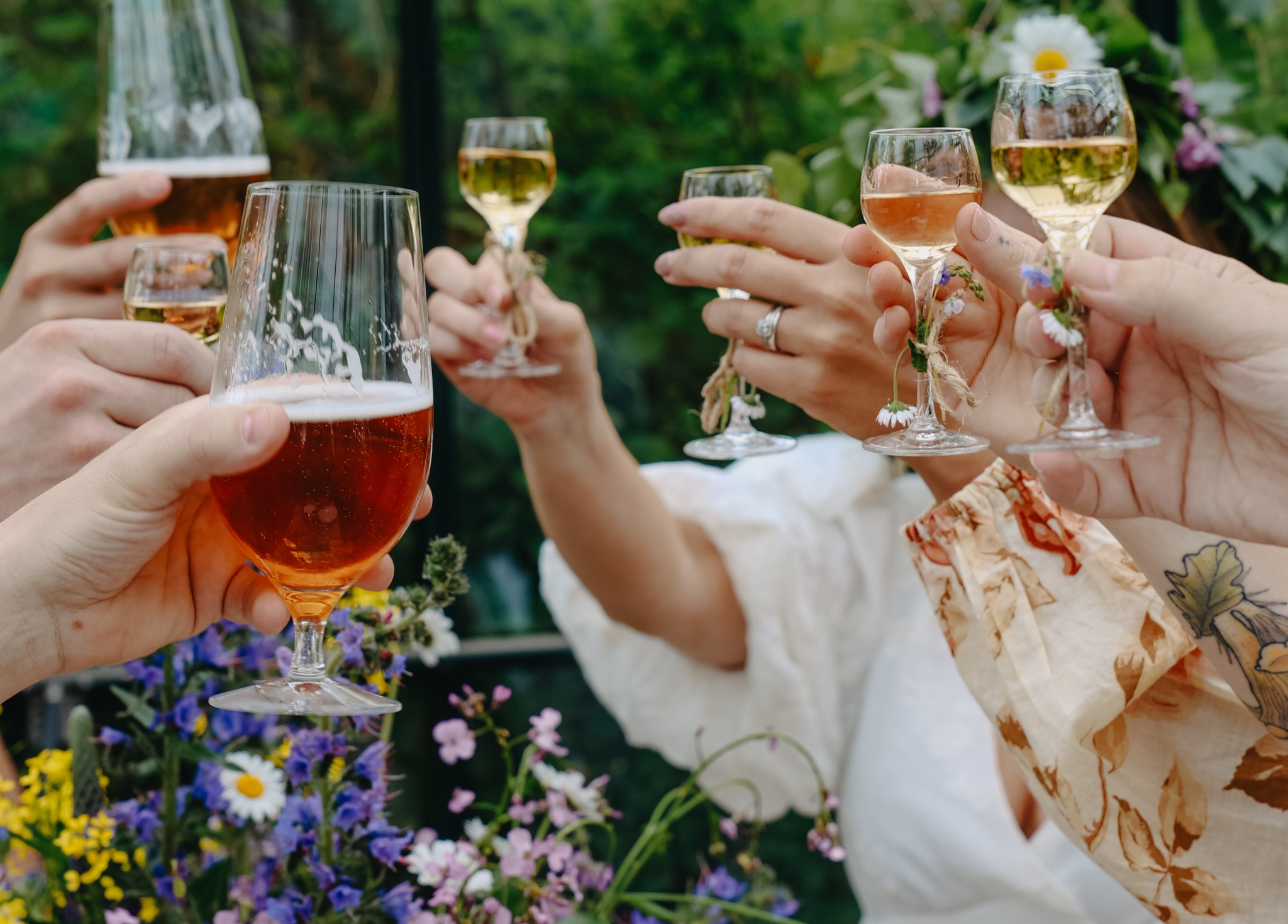 Venner skåler med øl og vin i en grønn hage omgitt av sommerblomster.