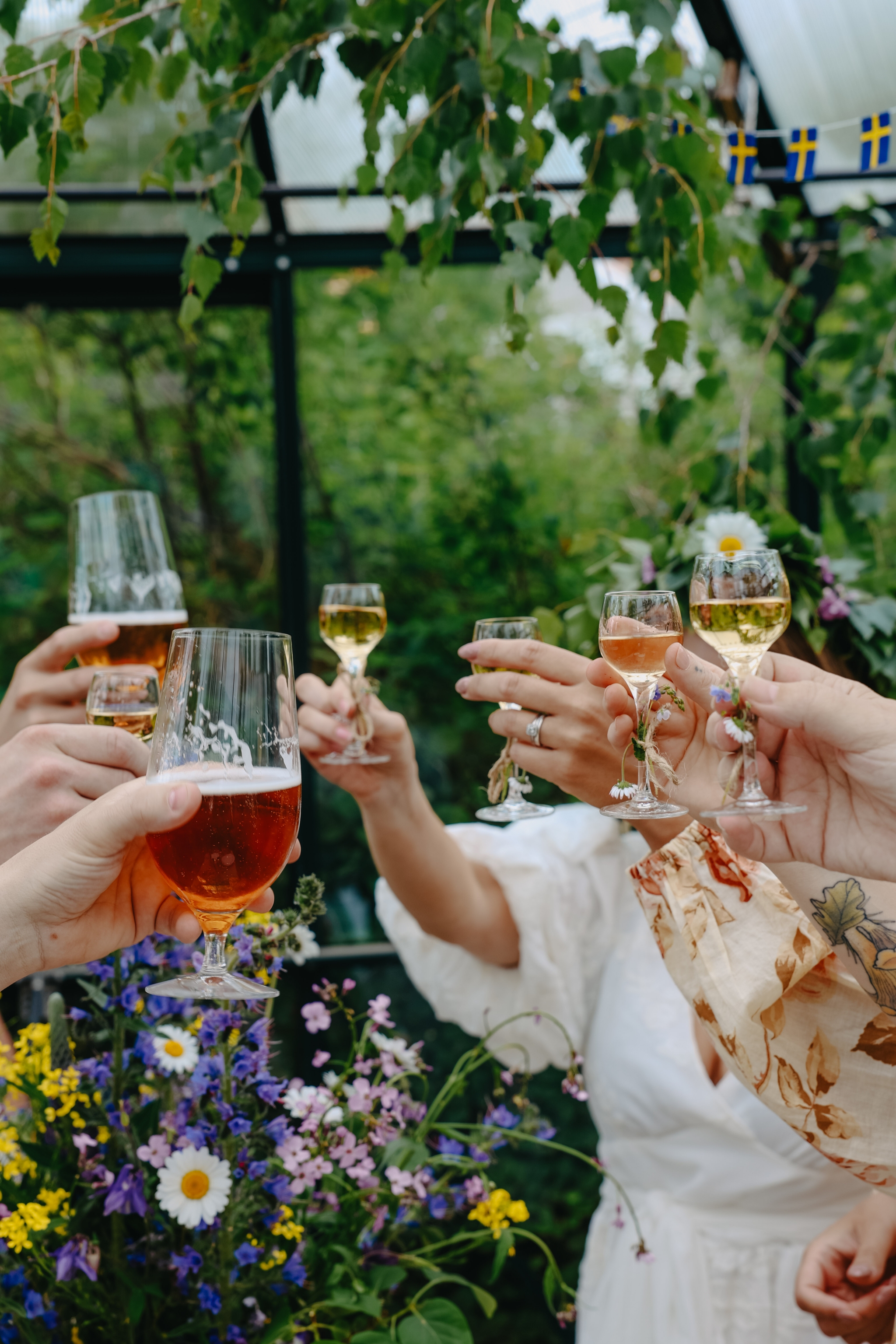 Vänner skålar med öl och vin under en somrig svensk fest omgiven av gröna blad och blommor.