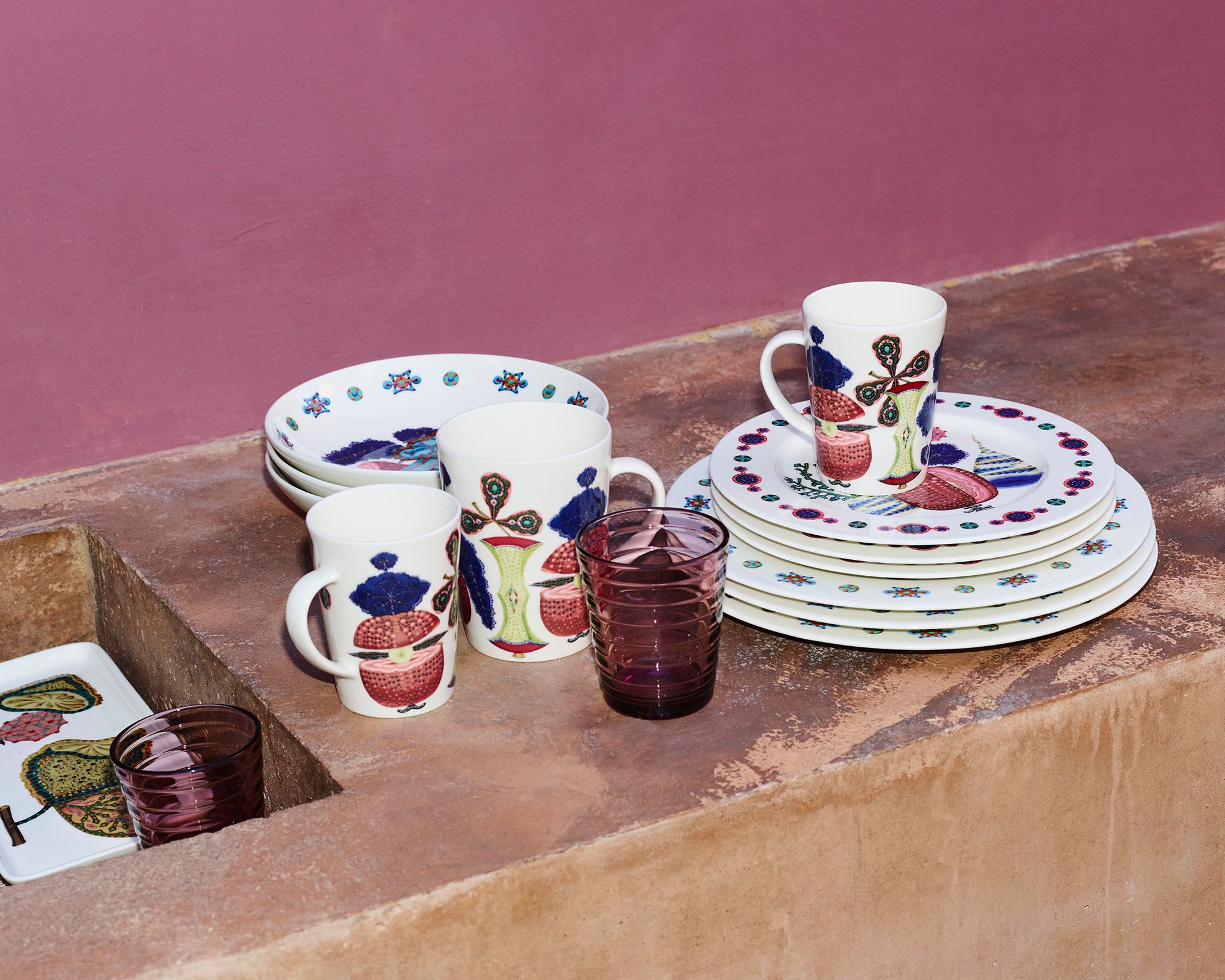 Valkoisia ruokailuvälineitä, joissa on värikkäitä hedelmäkuvioita, mukeja ja violetteja laseja aseteltuna teksturoidulle ruskealle pinnalle vaaleanpunaista seinää vasten.