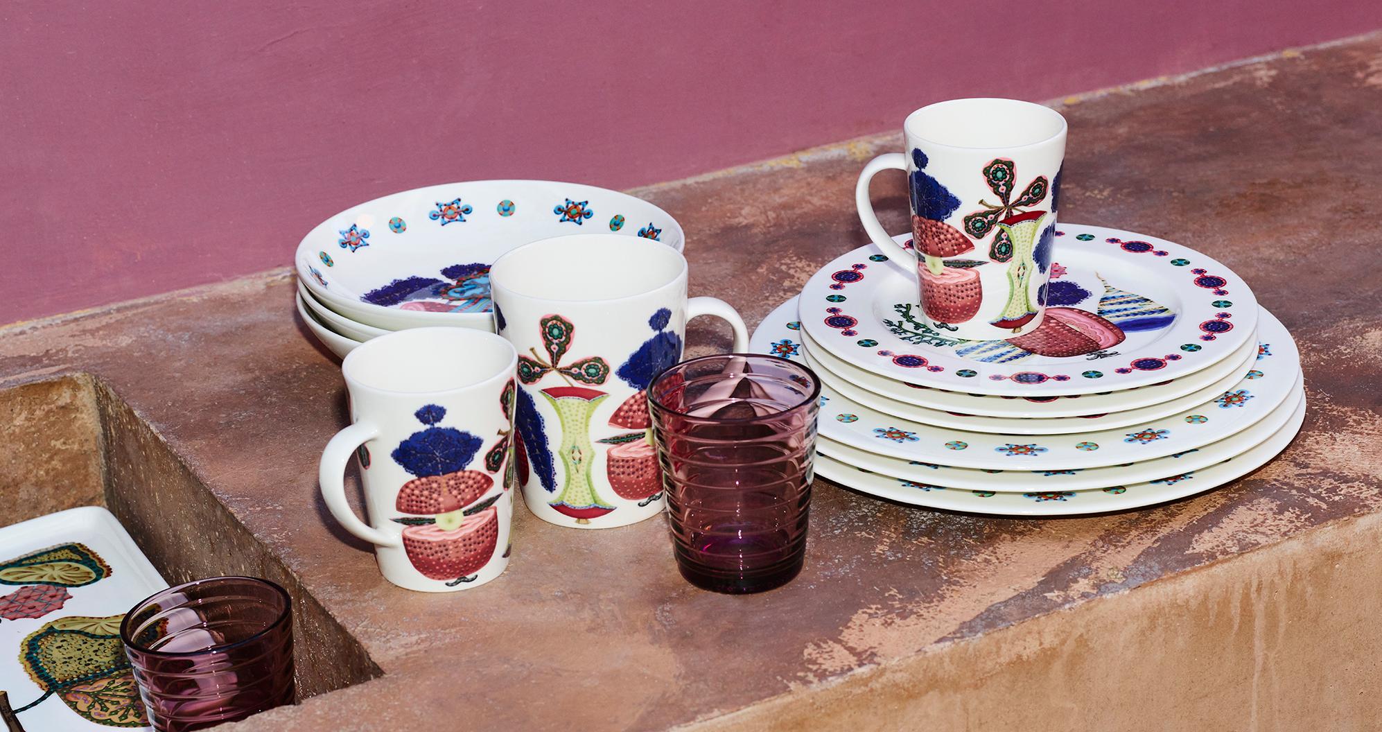 Valkoisia ruokailuvälineitä, joissa on värikkäitä hedelmäkuvioita, mukeja ja violetteja laseja aseteltuna teksturoidulle ruskealle pinnalle vaaleanpunaista seinää vasten.