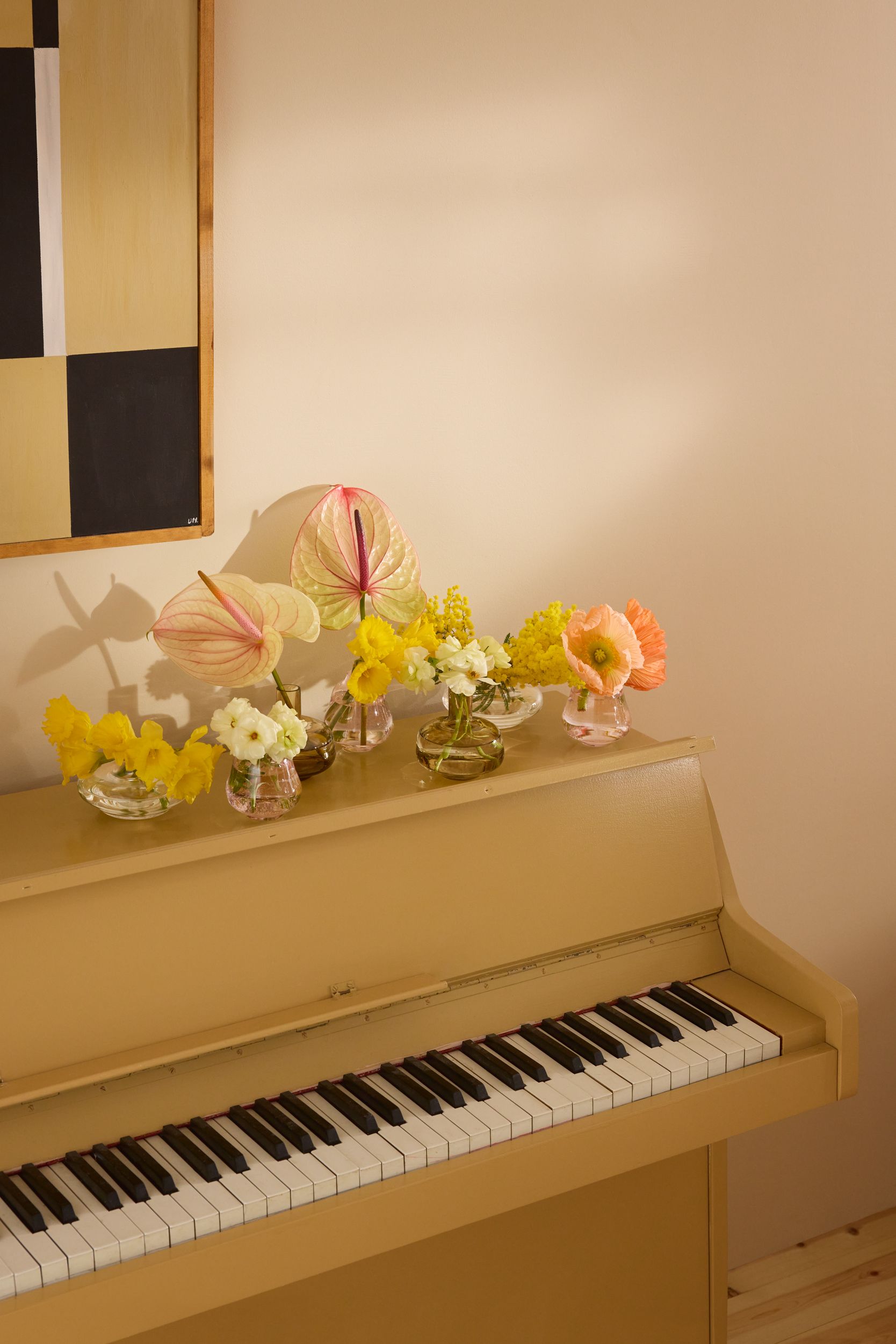 Ett ljusgult upprätt piano med flera små vaser ovanpå, fyllda med gula, rosa och orange blommor