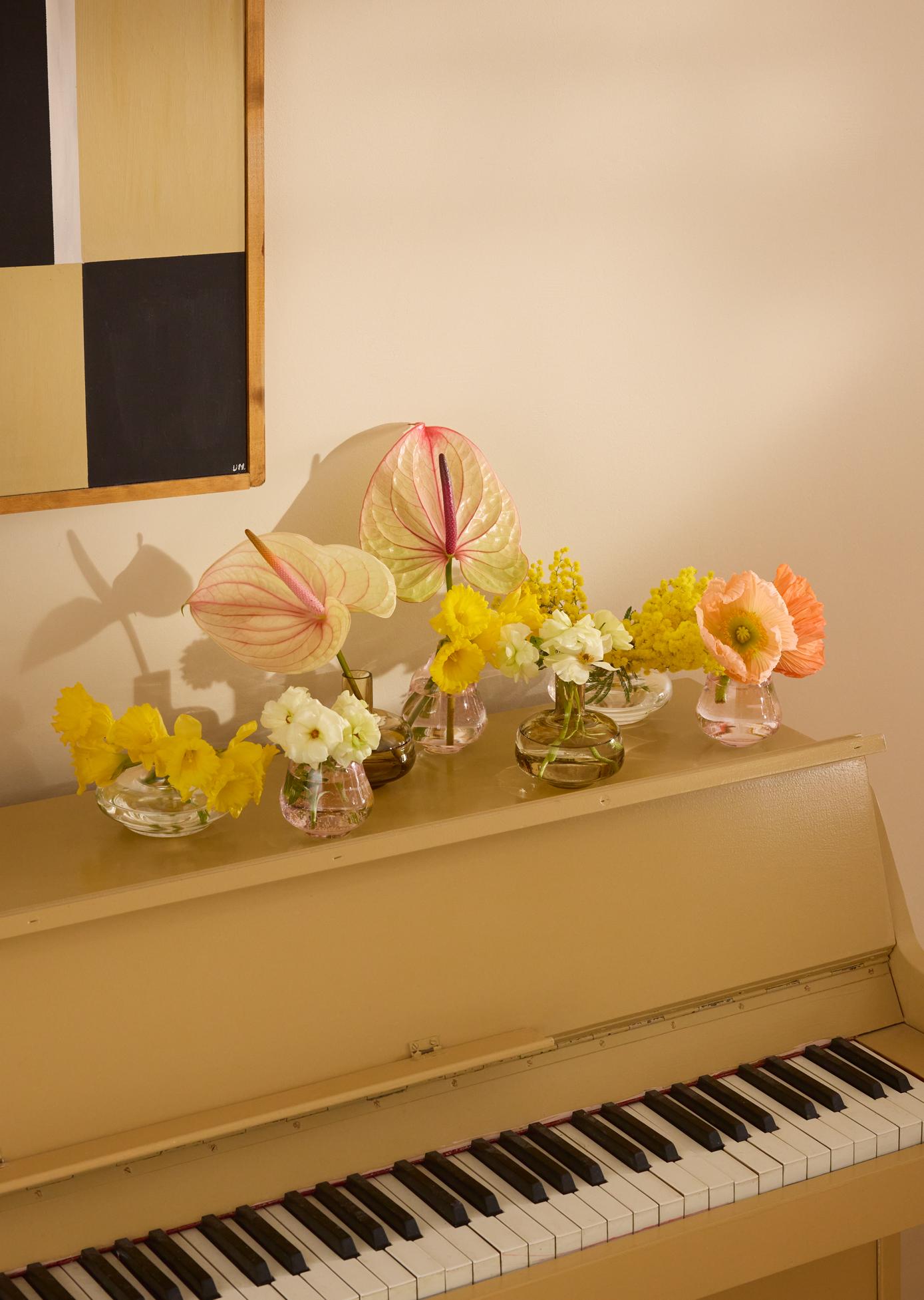 Et lysebrunt piano toppet med vaser med fargerike blomster, inkludert rosa anthurium, gule blomster og oransje valmuer