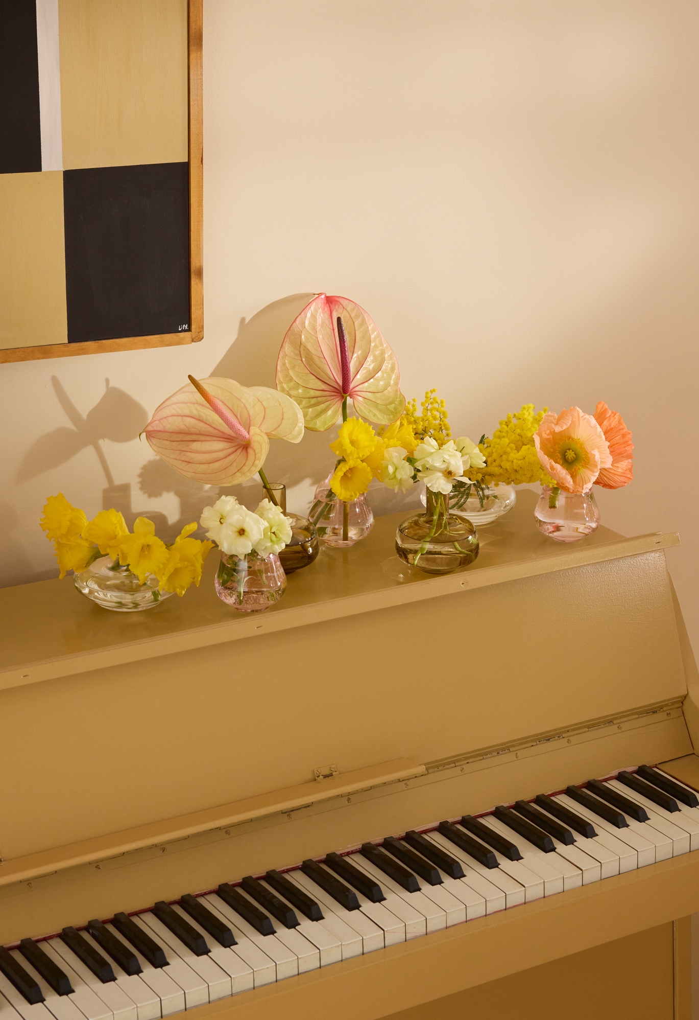 Et lysebrunt piano toppet med vaser med fargerike blomster, inkludert rosa anthurium, gule blomster og oransje valmuer