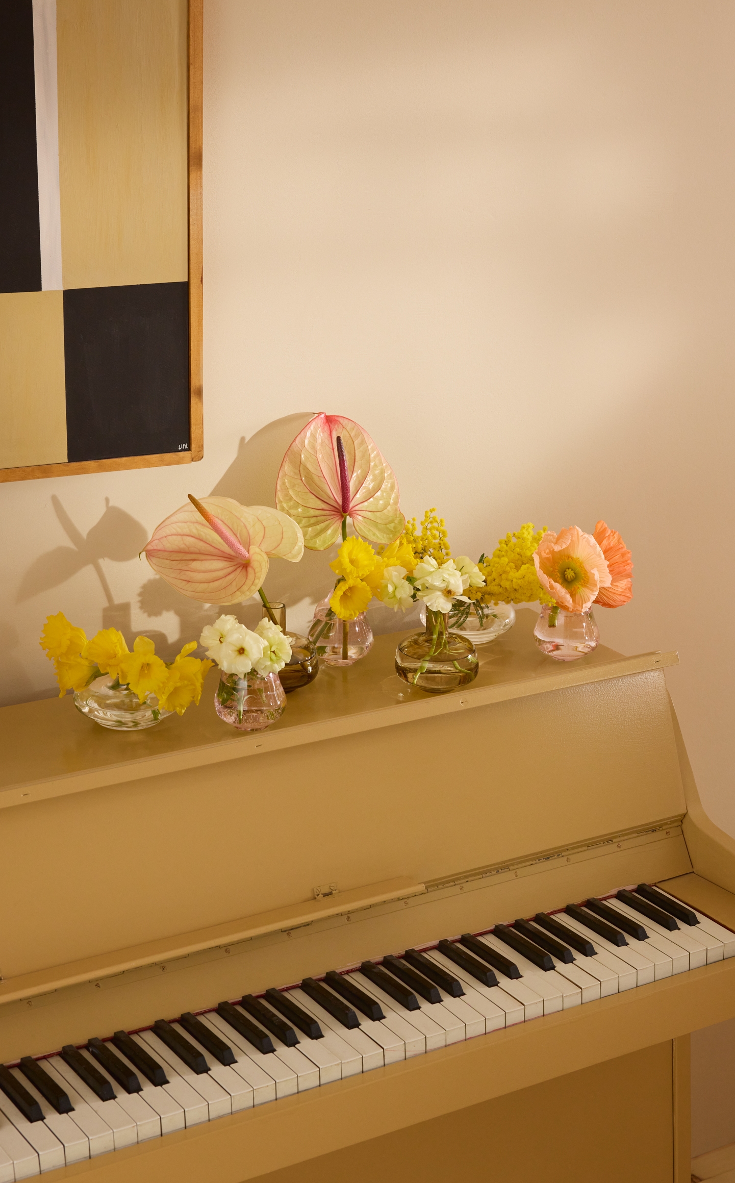 Ett ljusgult upprätt piano med flera små vaser ovanpå, fyllda med gula, rosa och orange blommor