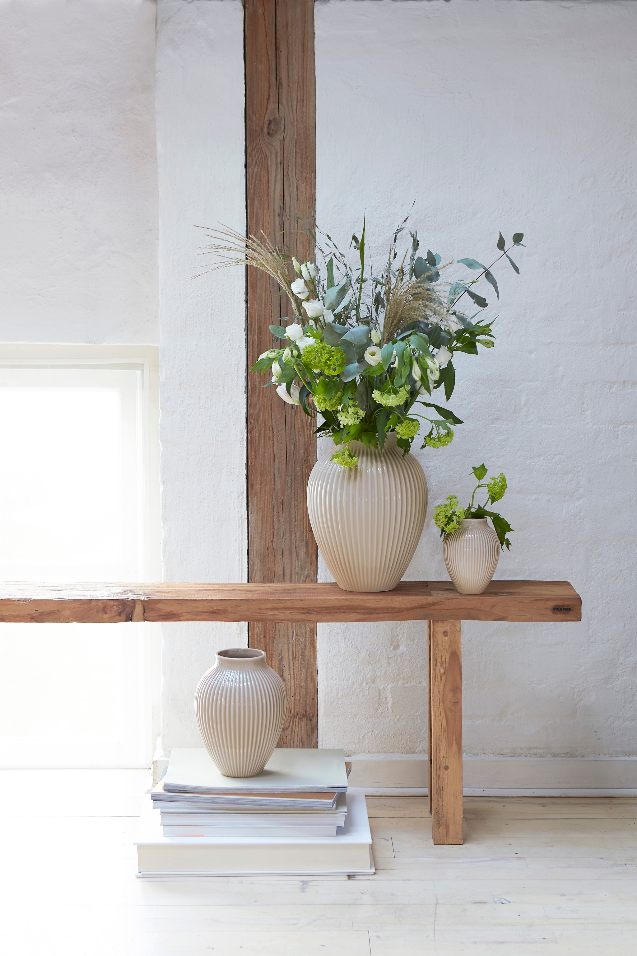 Lys og minimalistisk interiør med beige riflete vaser og grønne blomster på en enkel trebenk mot hvit murvegg.