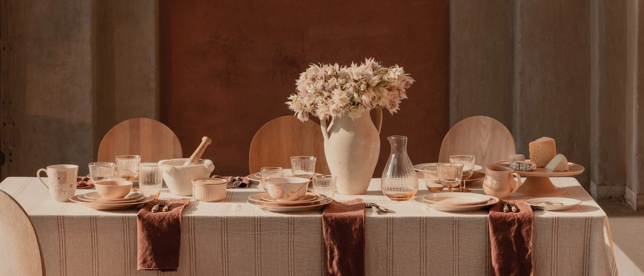 Et spisebord dekket med karamellfarget porselen, en blomstervase og trestoler, i et solfylt rom med steinsøyler og en rustfarget vegg.