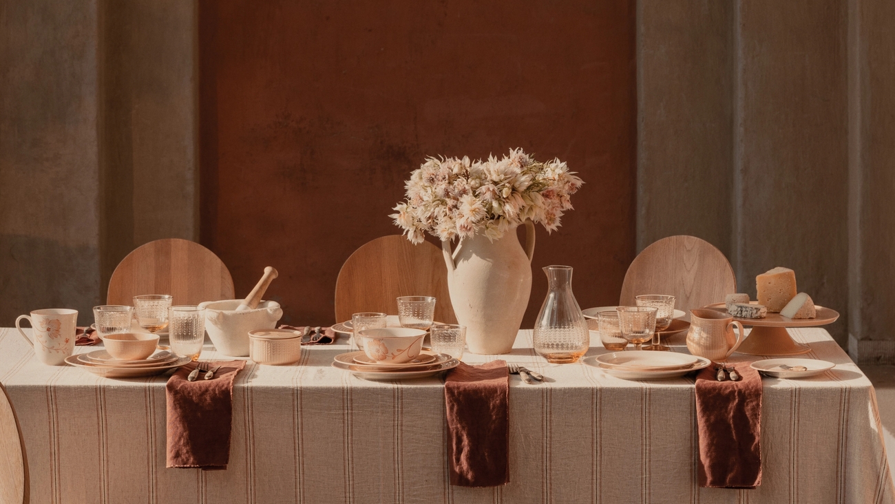 Et spisebord dekket med karamellfarget porselen, en blomstervase og trestoler, i et solfylt rom med steinsøyler og en rustfarget vegg.