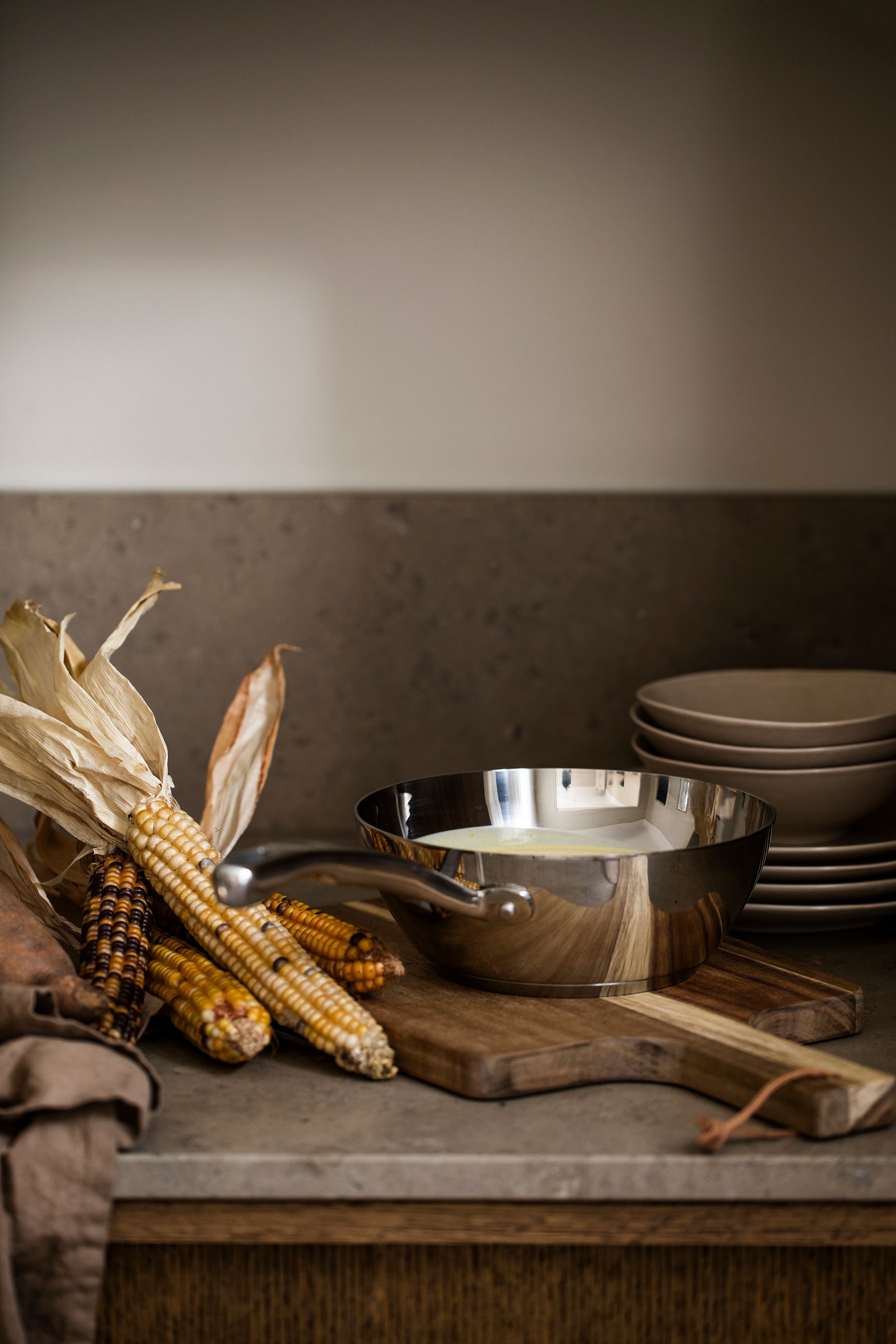 Dried corn cobs, a saucepan with liquid on a cutting board, and stacked dishes on a kitchen counter.