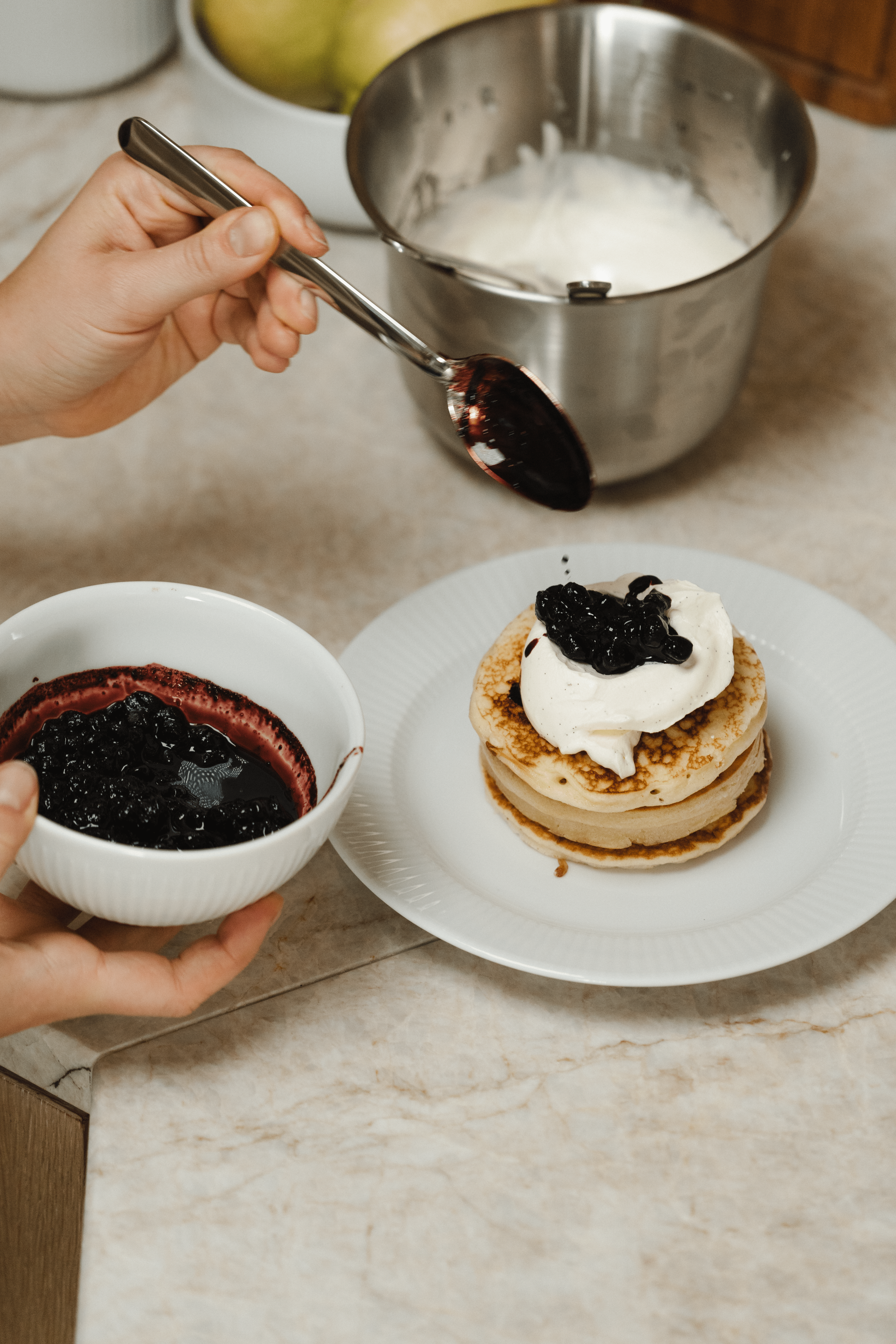 Person toppar en hög med pannkakor med vispgrädde och blåbärssylt i ett kök.