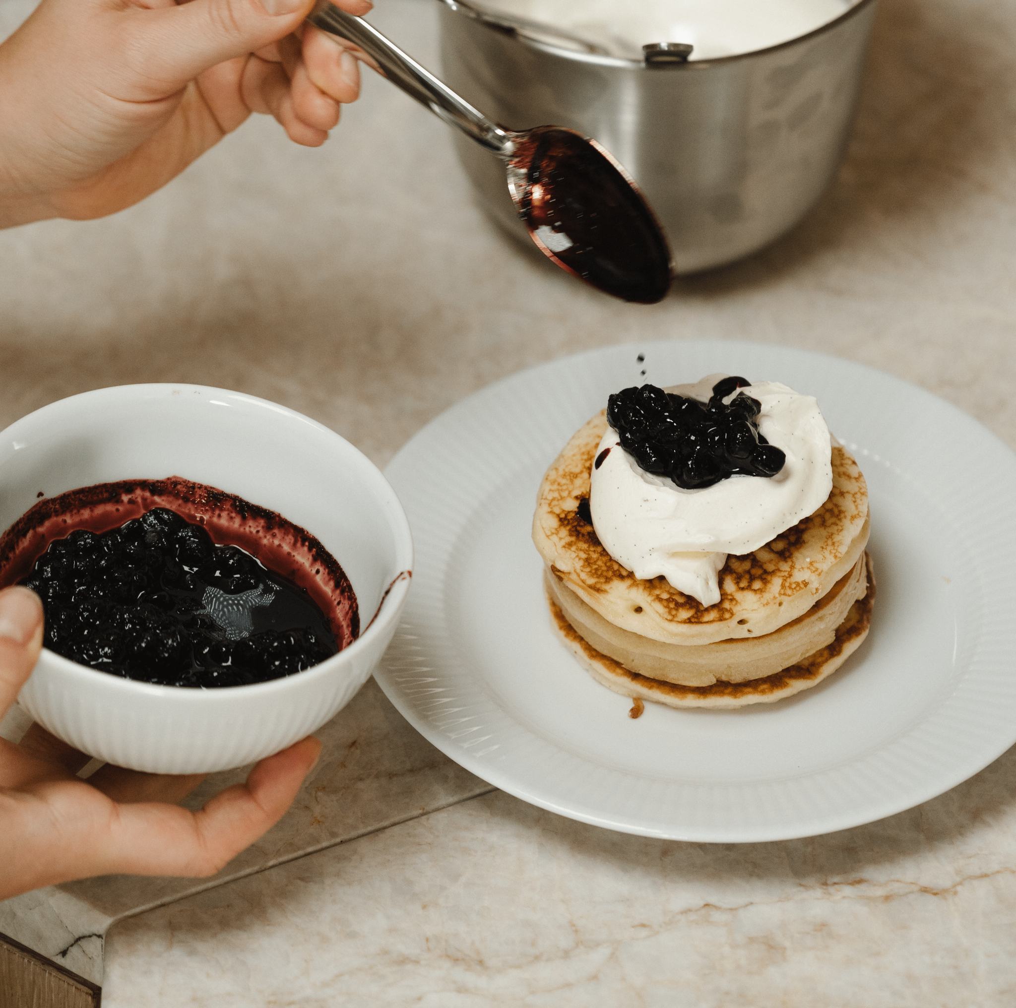 Person toppar en hög pannkakor med vispgrädde och blåbärssylt i ett ljust kök.