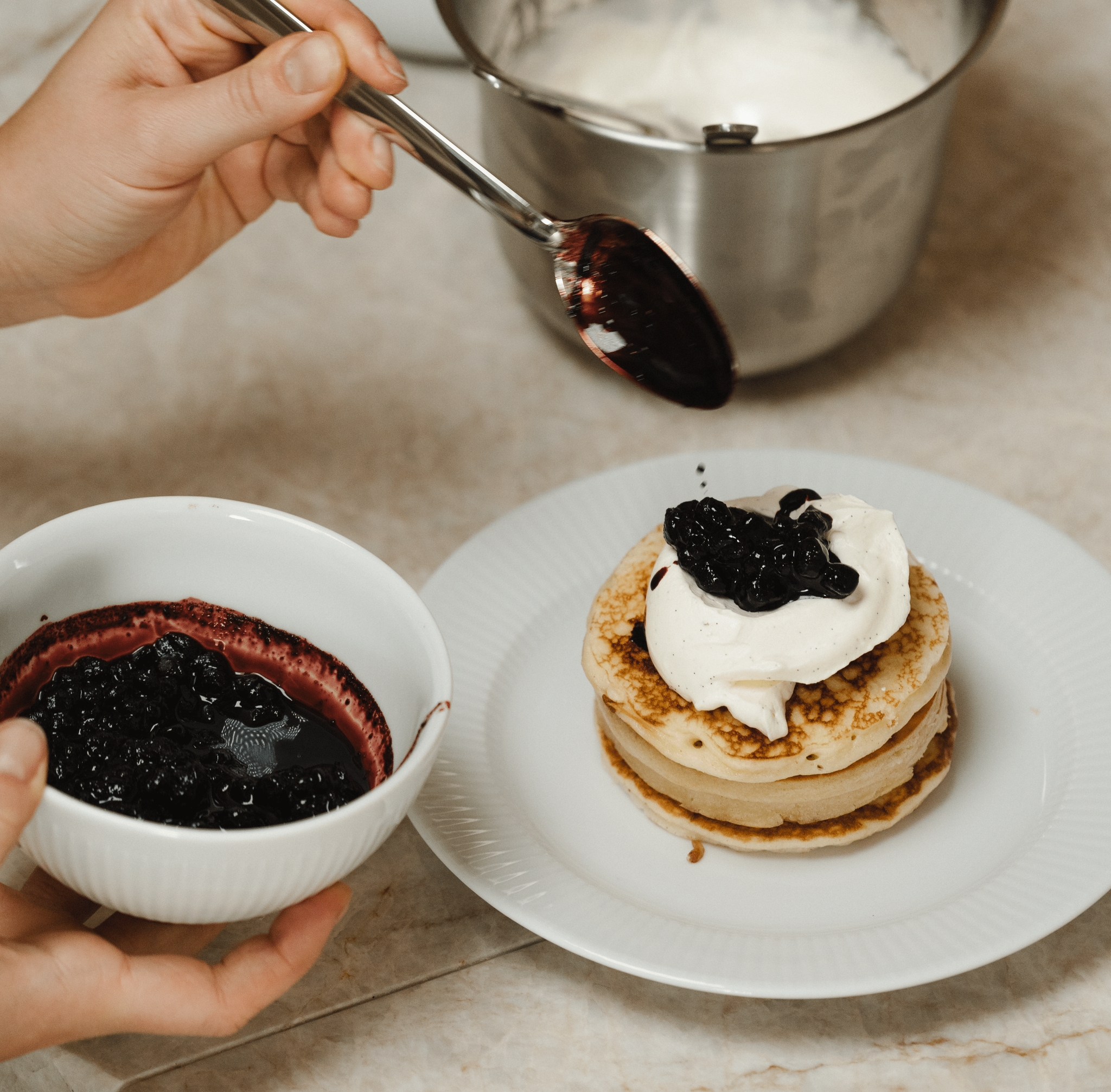 Person toppar en hög med pannkakor med vispgrädde och blåbärssylt i ett kök.