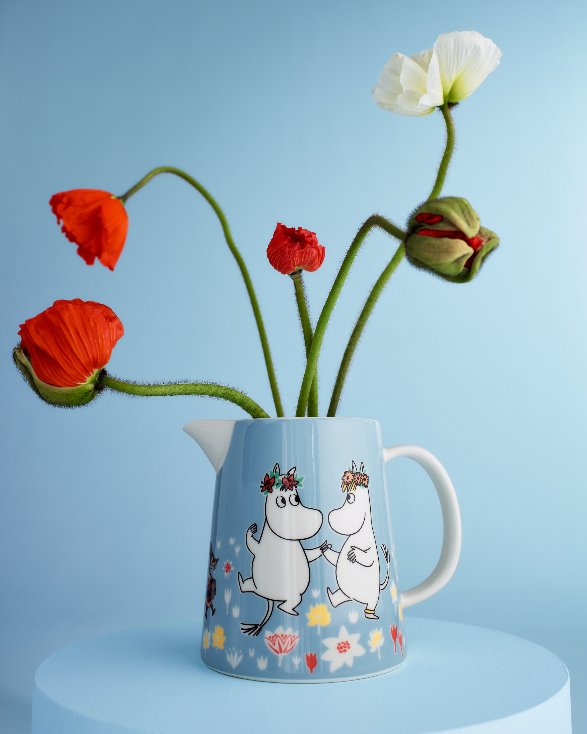 Blå Mumin-kanna med dansande Muminfigurer används som vas för röda och vita vallmoblommor mot ljusblå bakgrund.