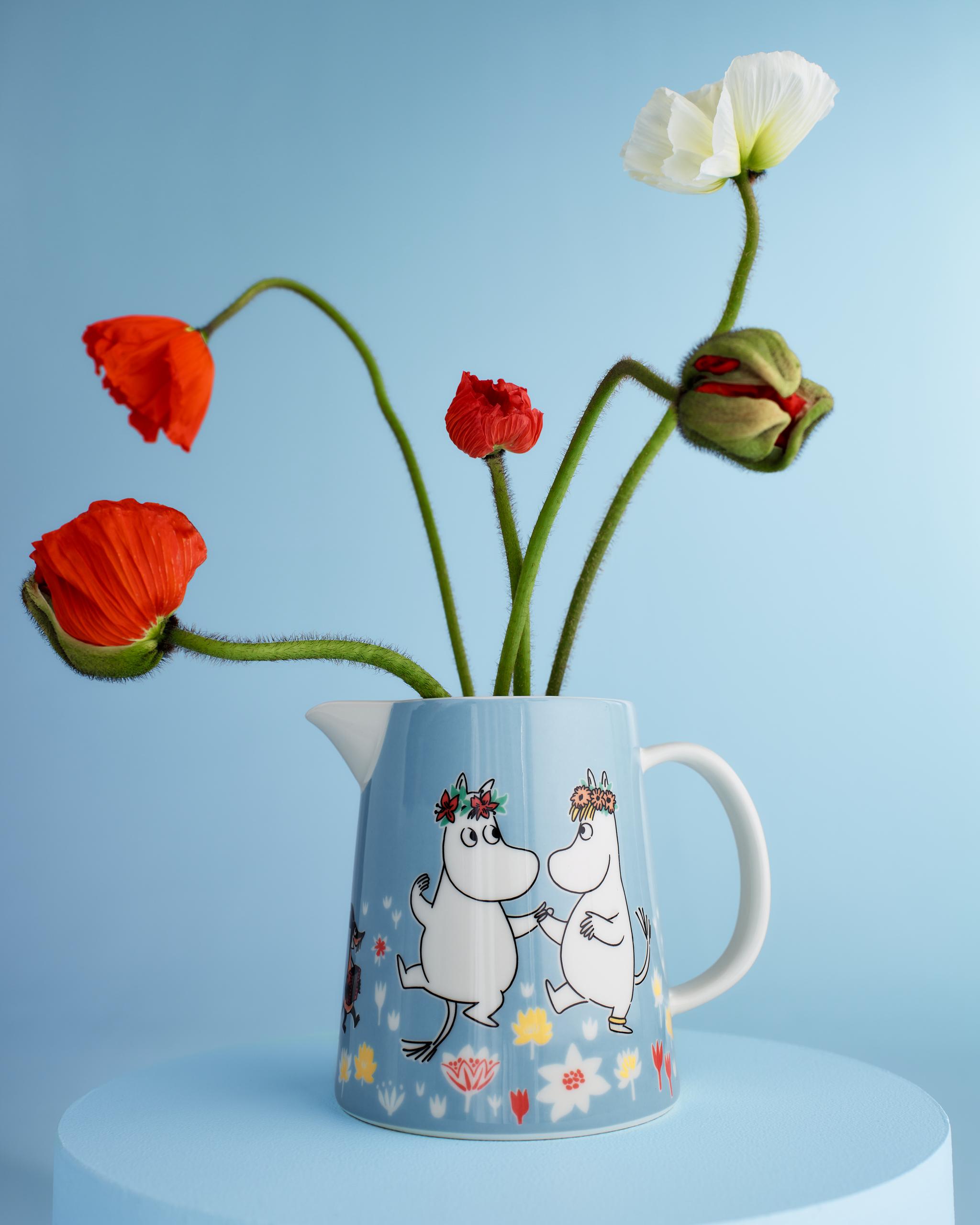 Blå Mumin-kanna med röda och vita vallmoblommor mot ljusblå bakgrund.