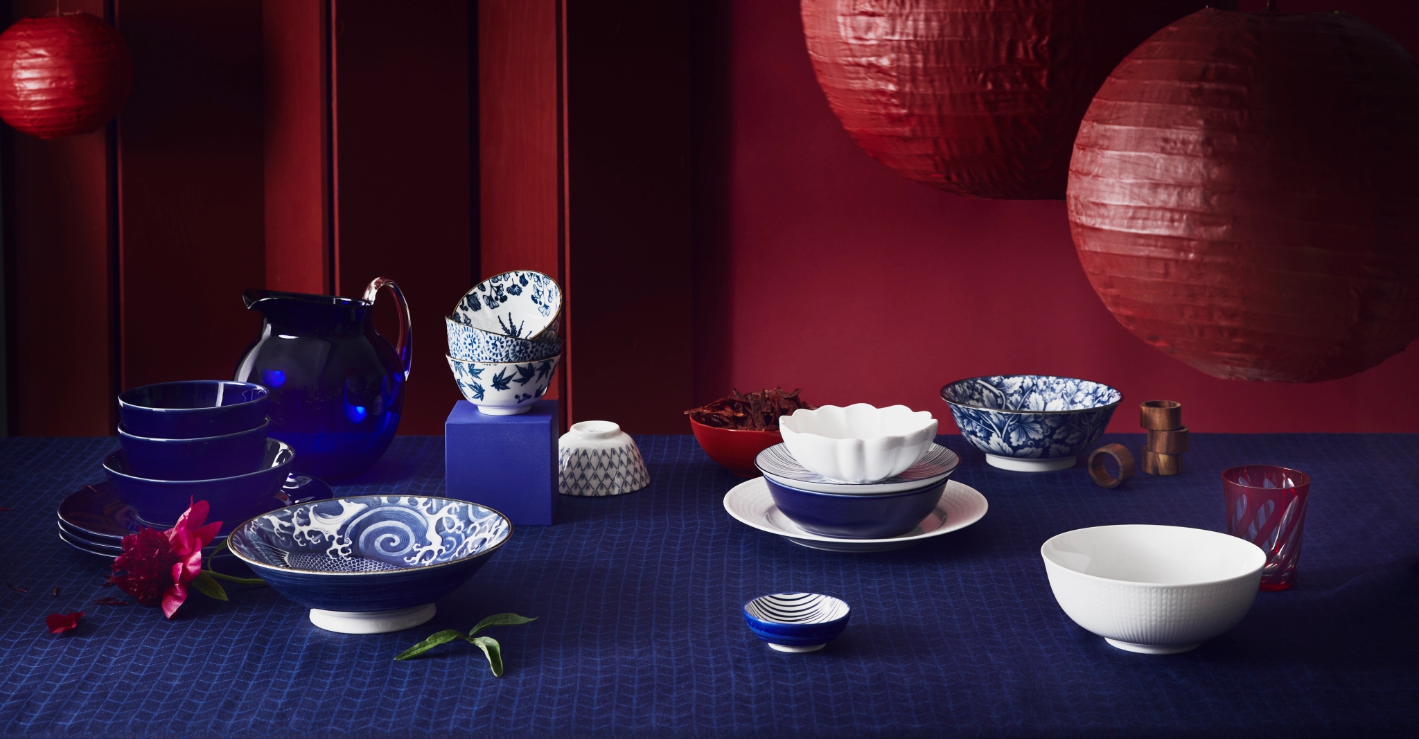 Blue, white, and patterned dinnerware on a blue tablecloth, featuring a pitcher, bowls, and plates, with a red background and two hanging red paper lanterns.