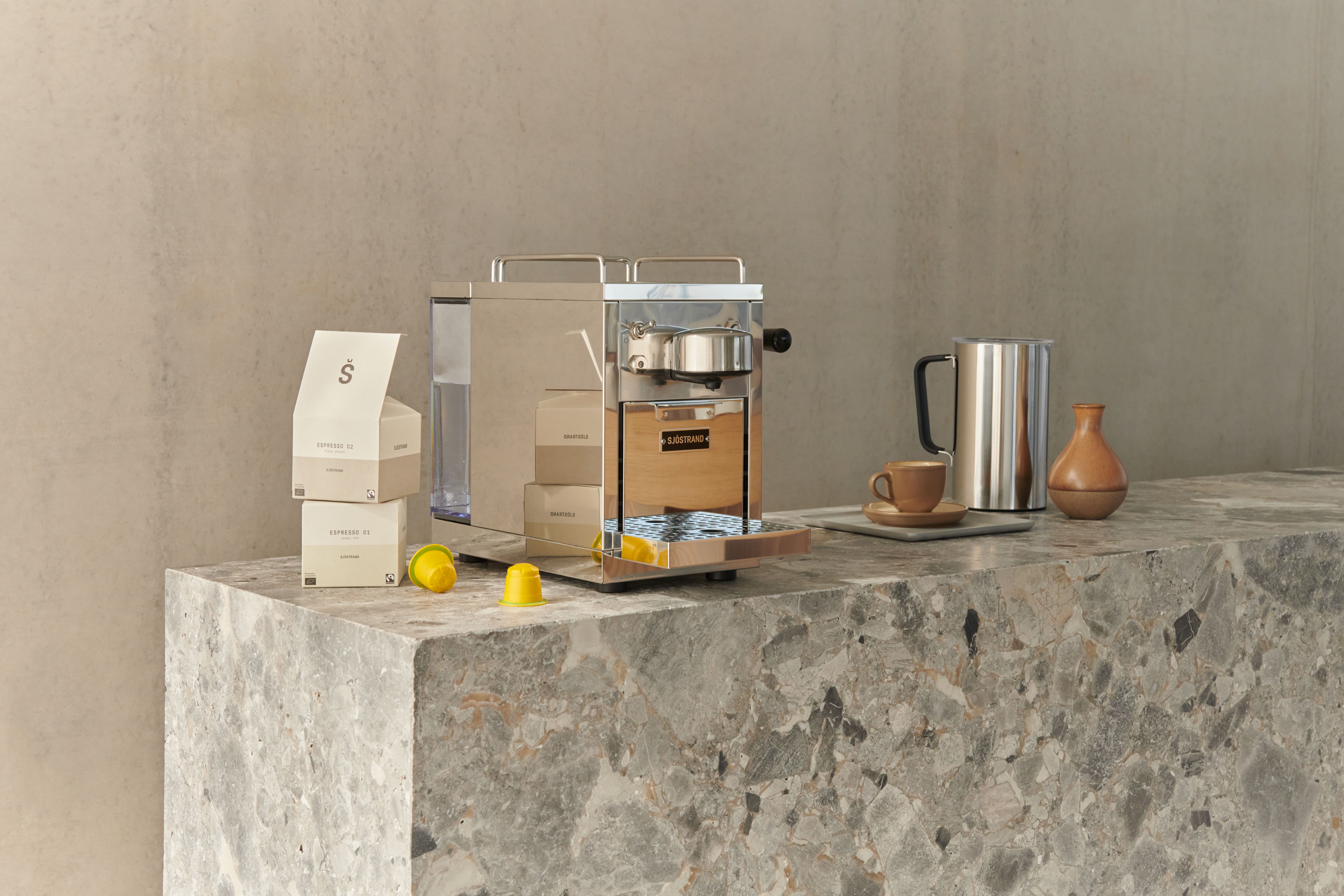 Metallinen kapselikeitin harmaalla marmoritiskillä, jossa on kahvipakkauksia, keltaisia kapseleita, metallinen kaadin ja keraaminen kuppi.